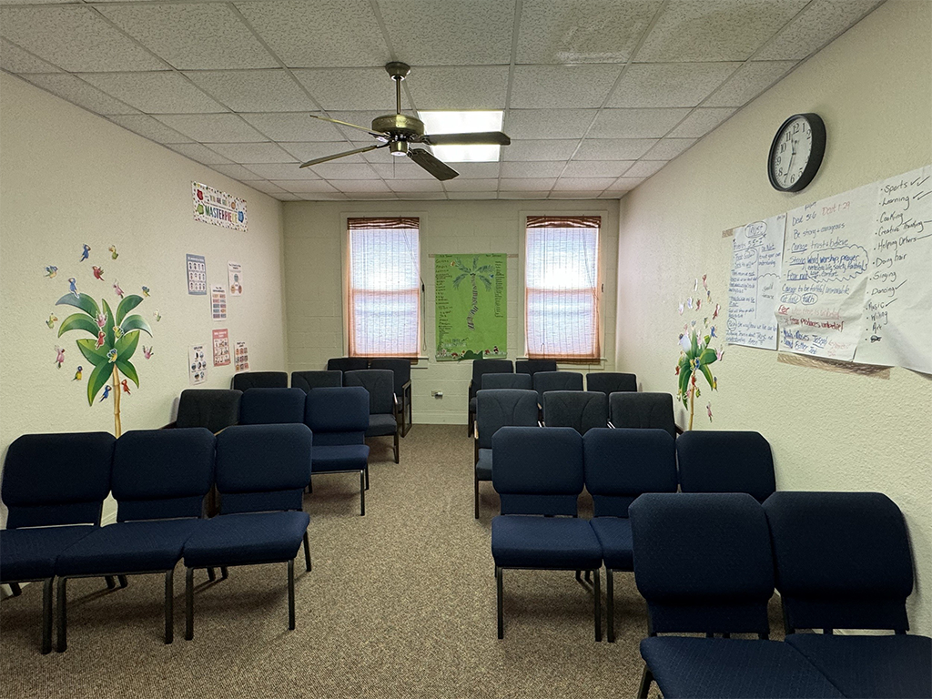 Classroom with rows of navy blue chairs, two windows with pink curtains, a ceiling fan, a whiteboard with handwritten notes, colorful wall decorations, and a clock showing 9:10.