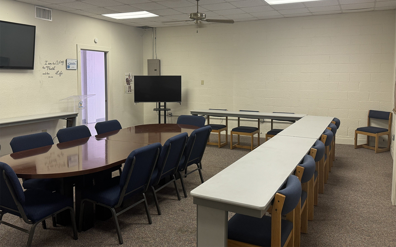 A conference room with a long wooden table surrounded by blue chairs and a white table with more chairs along the back wall. There is a large flat screen TV mounted on a stand and a smaller television on the wall. The room has a ceiling fan and a door leading outside, with some wall decorations.
