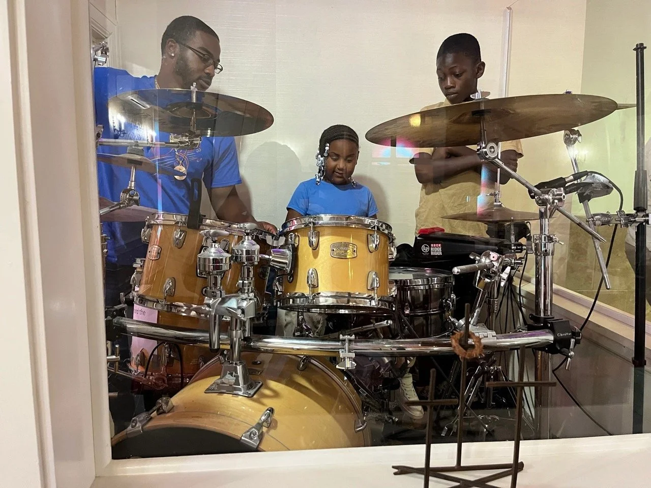 A man and a boy playing a drum set in a recording studio, with a young girl in blue observing.