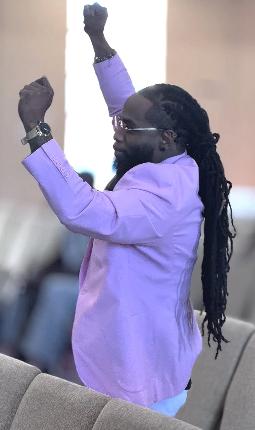 A man with long dreadlocks, wearing glasses and a purple blazer, is raising his arms in a gesture of celebration or empowerment.