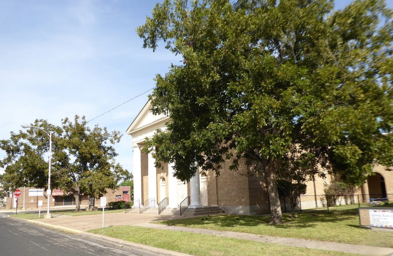 A church with white columns and brick walls next to a large green tree along a street with a stop sign.