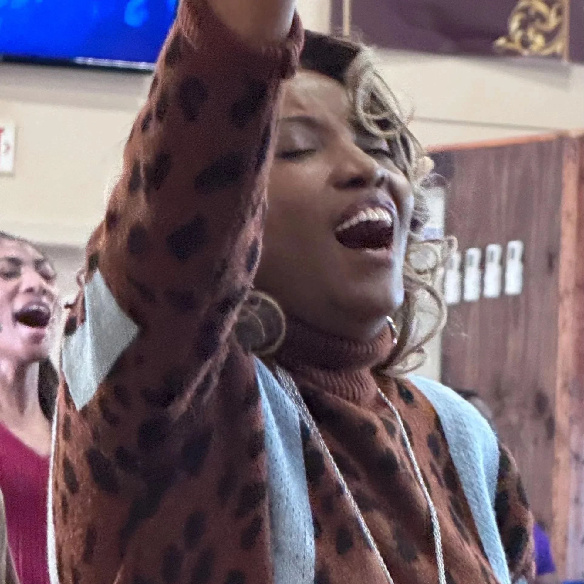 A woman with curly hair, wearing a brown and gray cardigan, is singing with her eyes closed and mouth open, in a lively setting.
