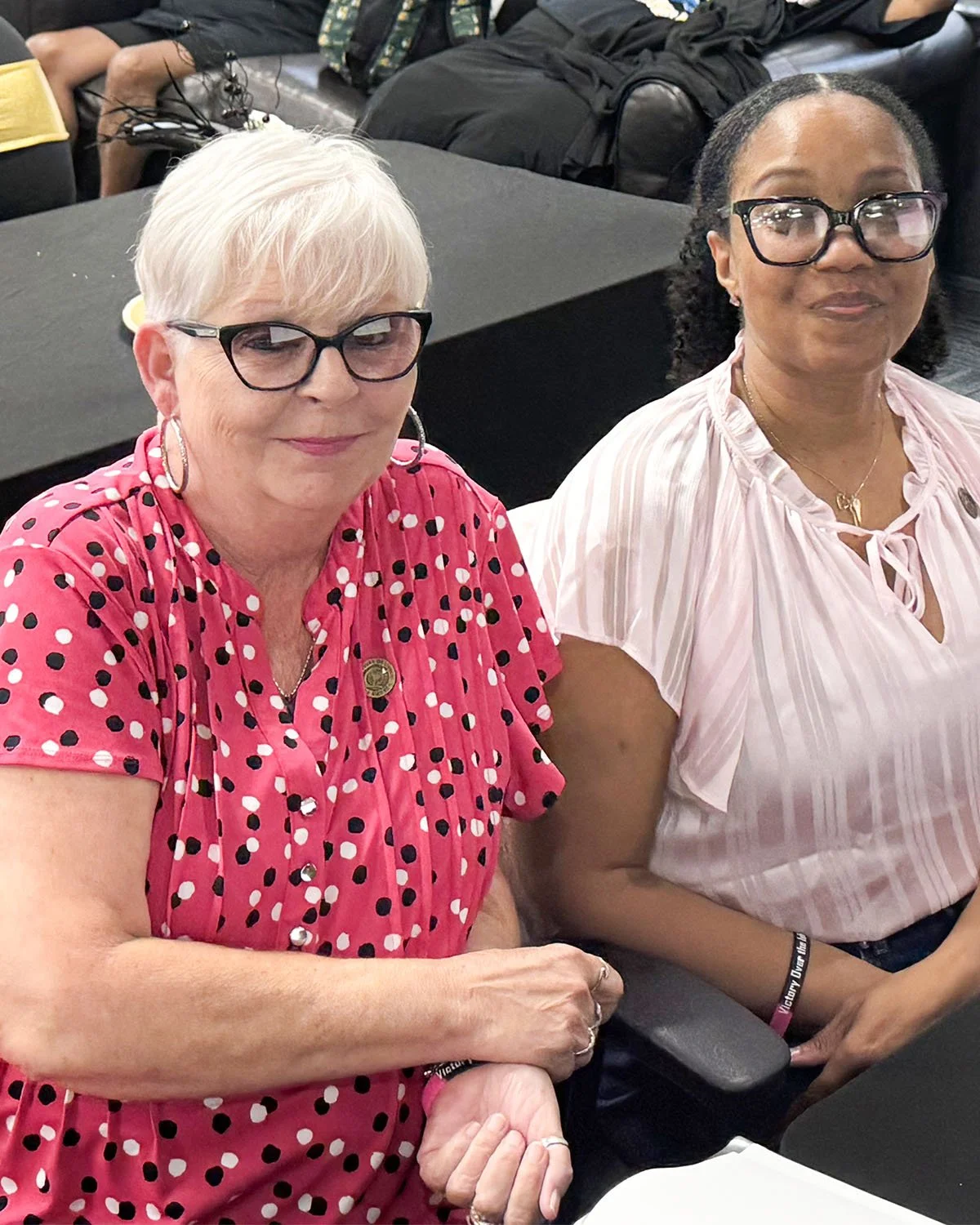 Two women sitting at a table at an indoor event, smiling, wearing glasses, with people and belongings in the background.