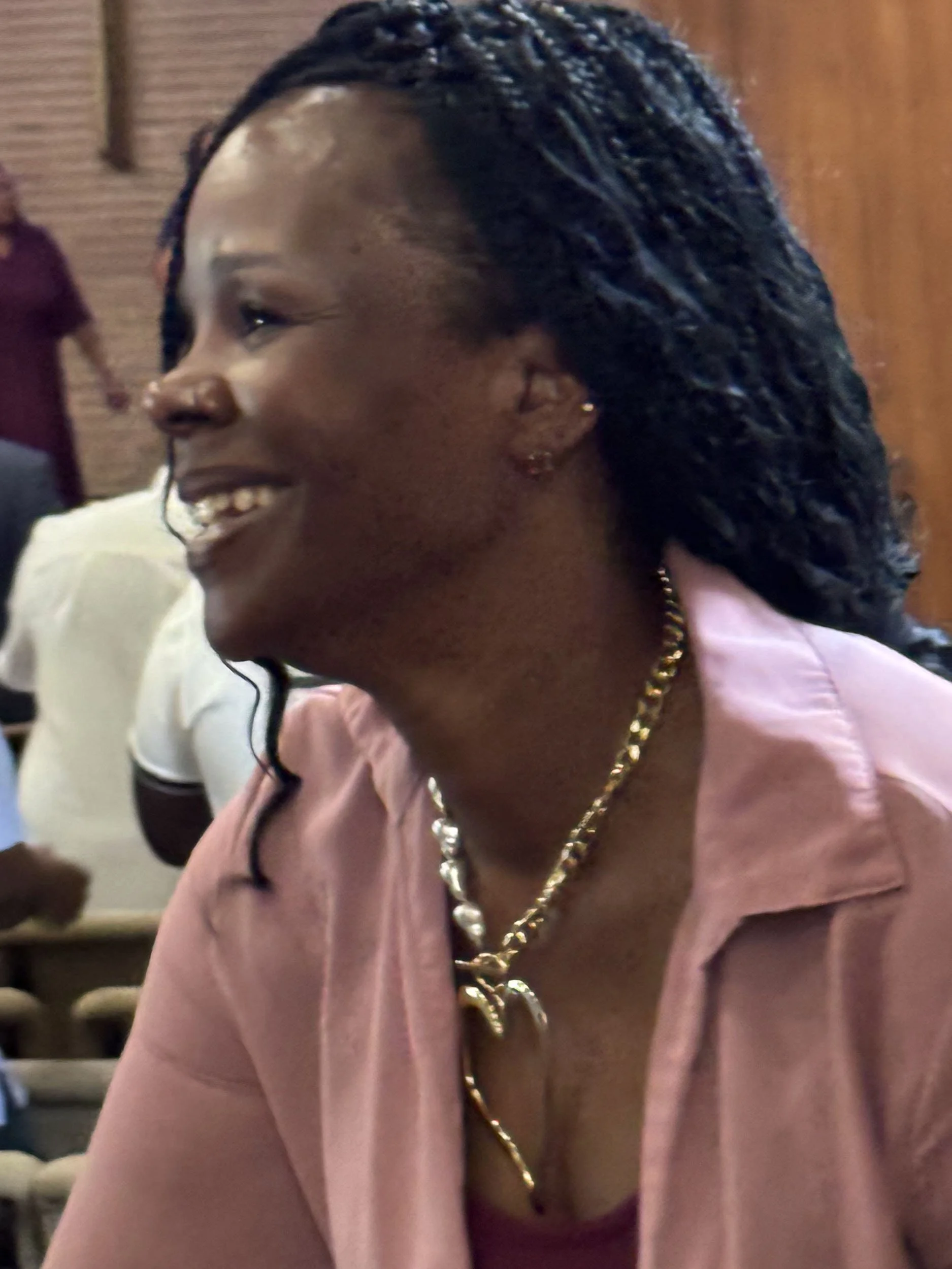 A smiling woman with braided hair wearing a pink shirt and gold jewelry, enjoying a moment at a social gathering.