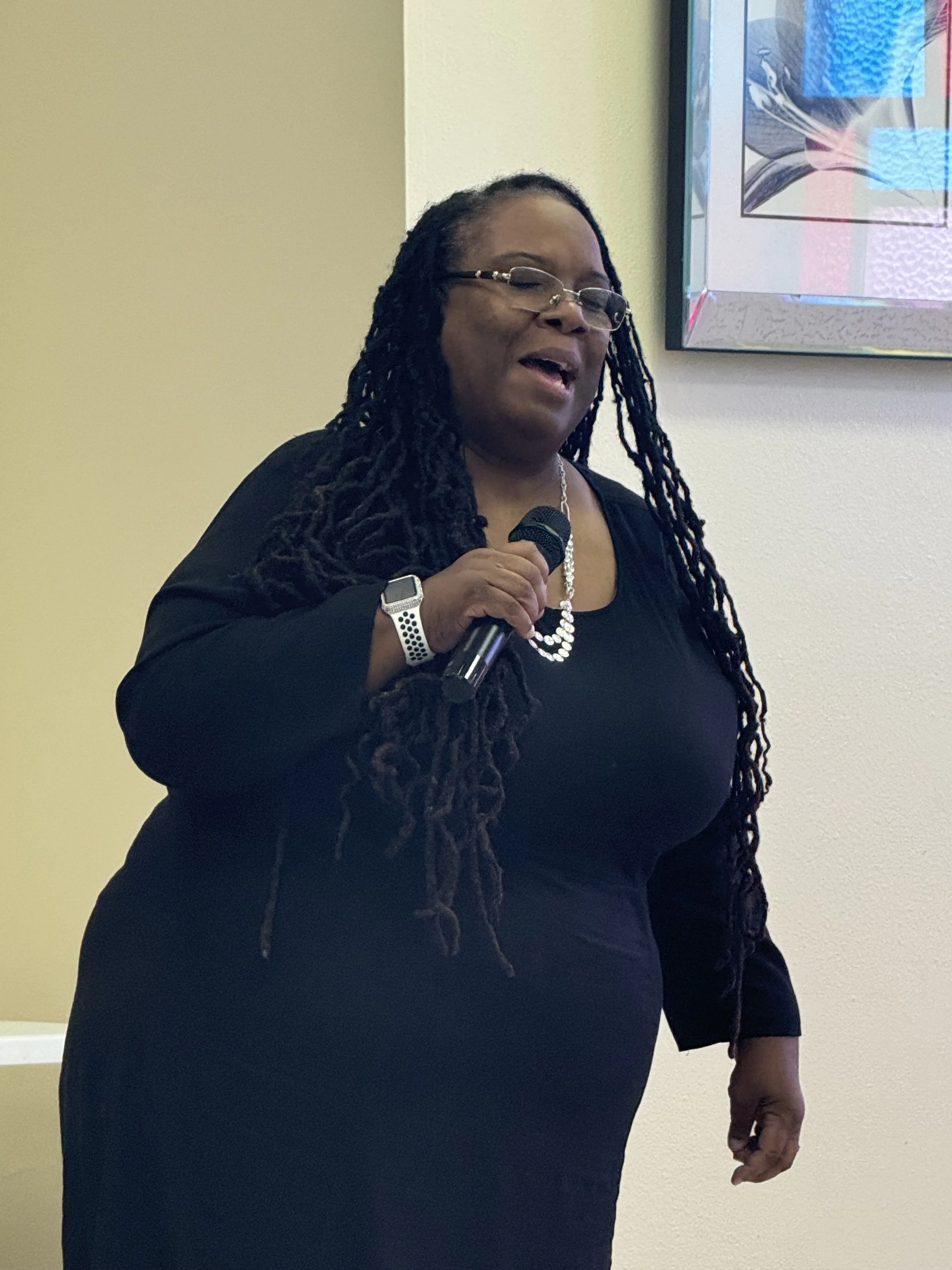A woman with long black dreadlocks, glasses, and a pearl necklace, holding a microphone and singing or speaking, dressed in a black outfit, standing indoors in front of a light-colored wall and a framed artwork.