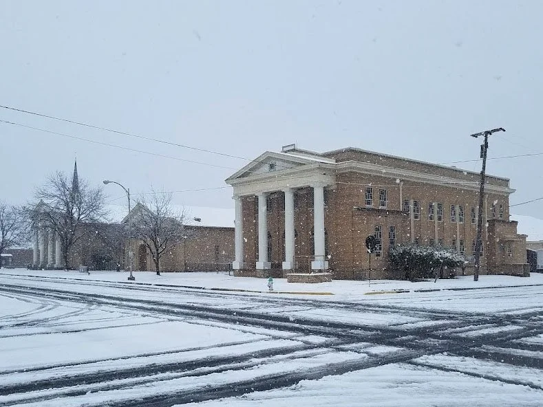 A snowy scene with a large building featuring classical architecture, including four large pillars at the entrance, on a winter day with snow covering the ground and snowflakes falling.