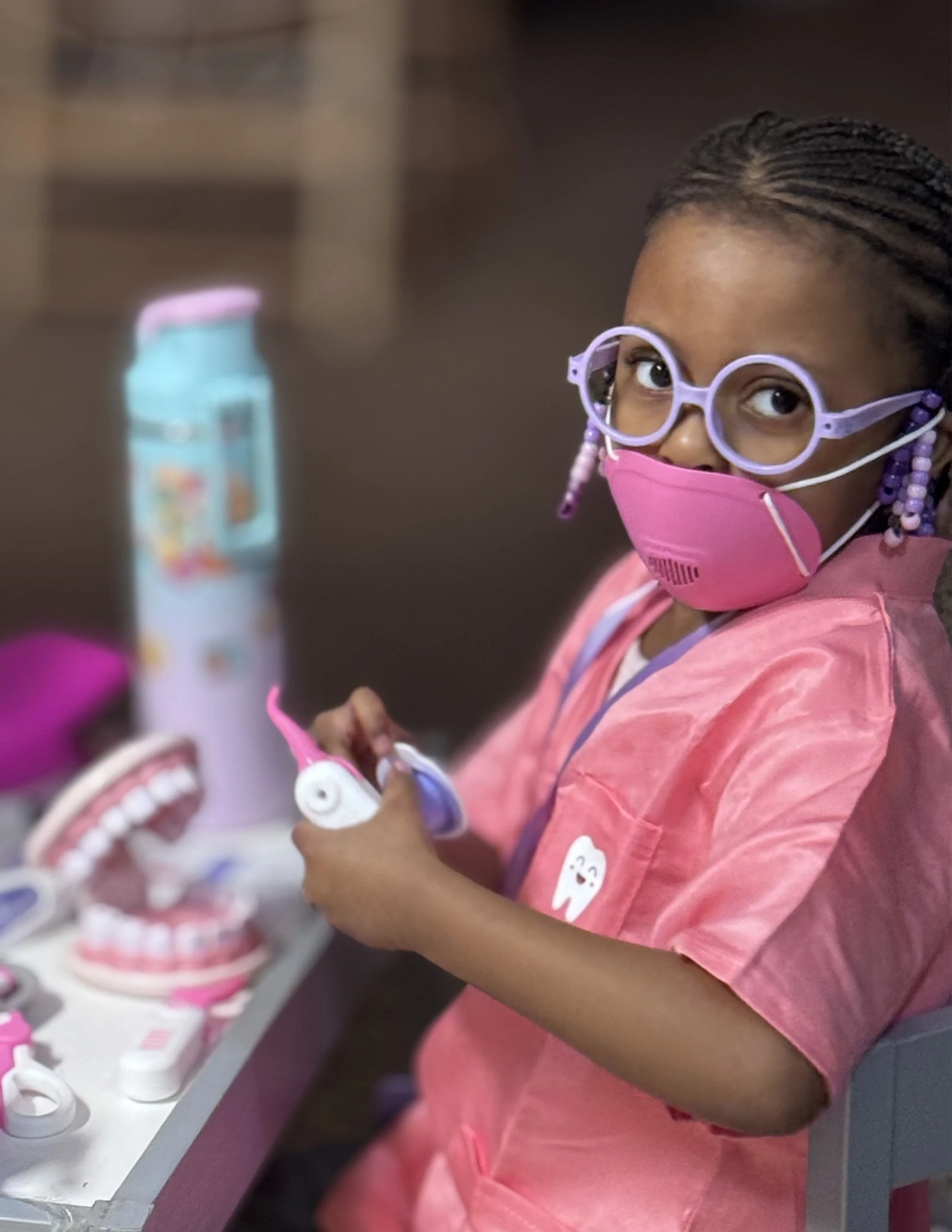 Young girl wearing a pink mask, purple glasses, and a pink dental apron, playing with a toy dental kit at a table with a large model of teeth, dental tools, and a water bottle in the background.