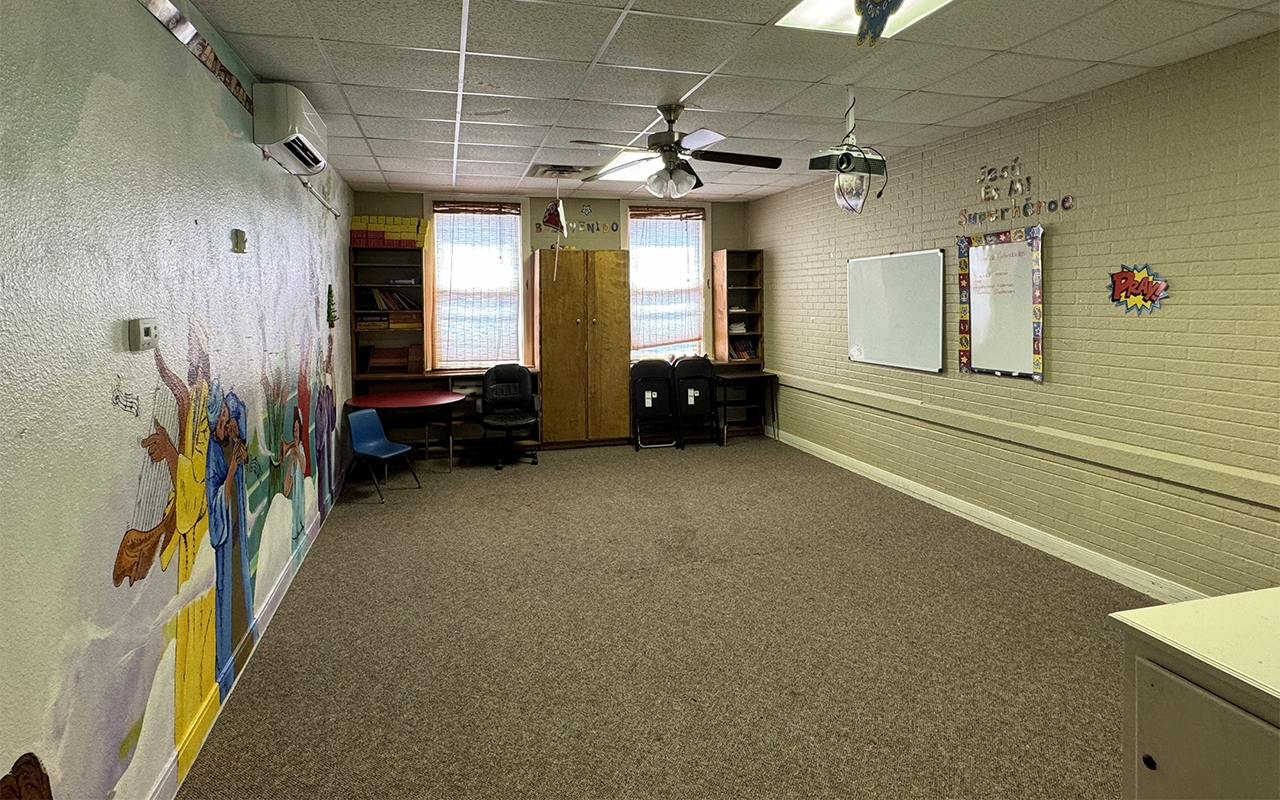 Large classroom with a painted wall mural of Jesus Christ and children, windows with blinds, chairs against the wall, a whiteboard on the wall, and various decorations including a projector and signs.