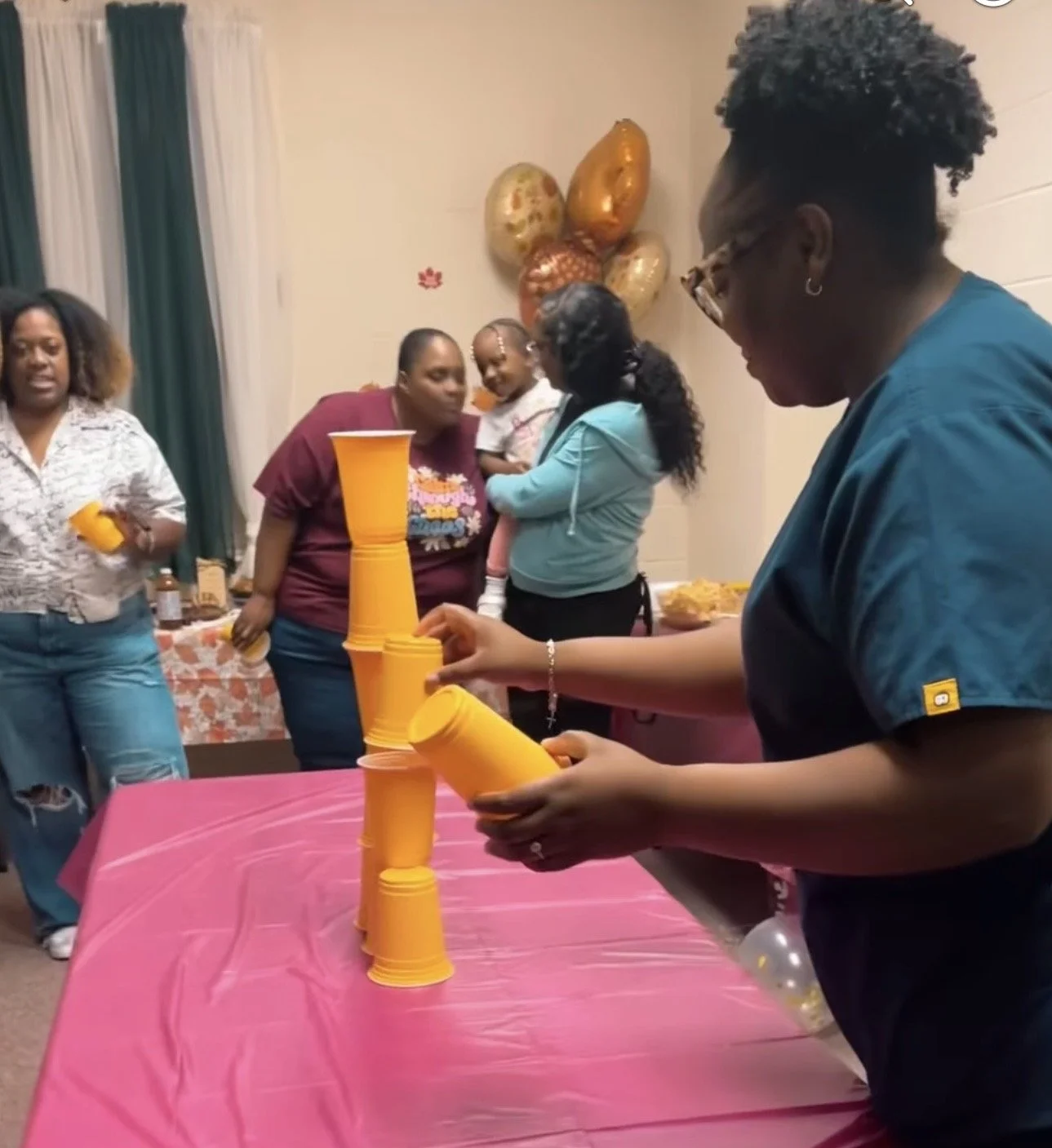 A woman is stacking yellow cups on a pink tablecloth at a family gathering, while other women and a child observe nearby, with balloons in the background.