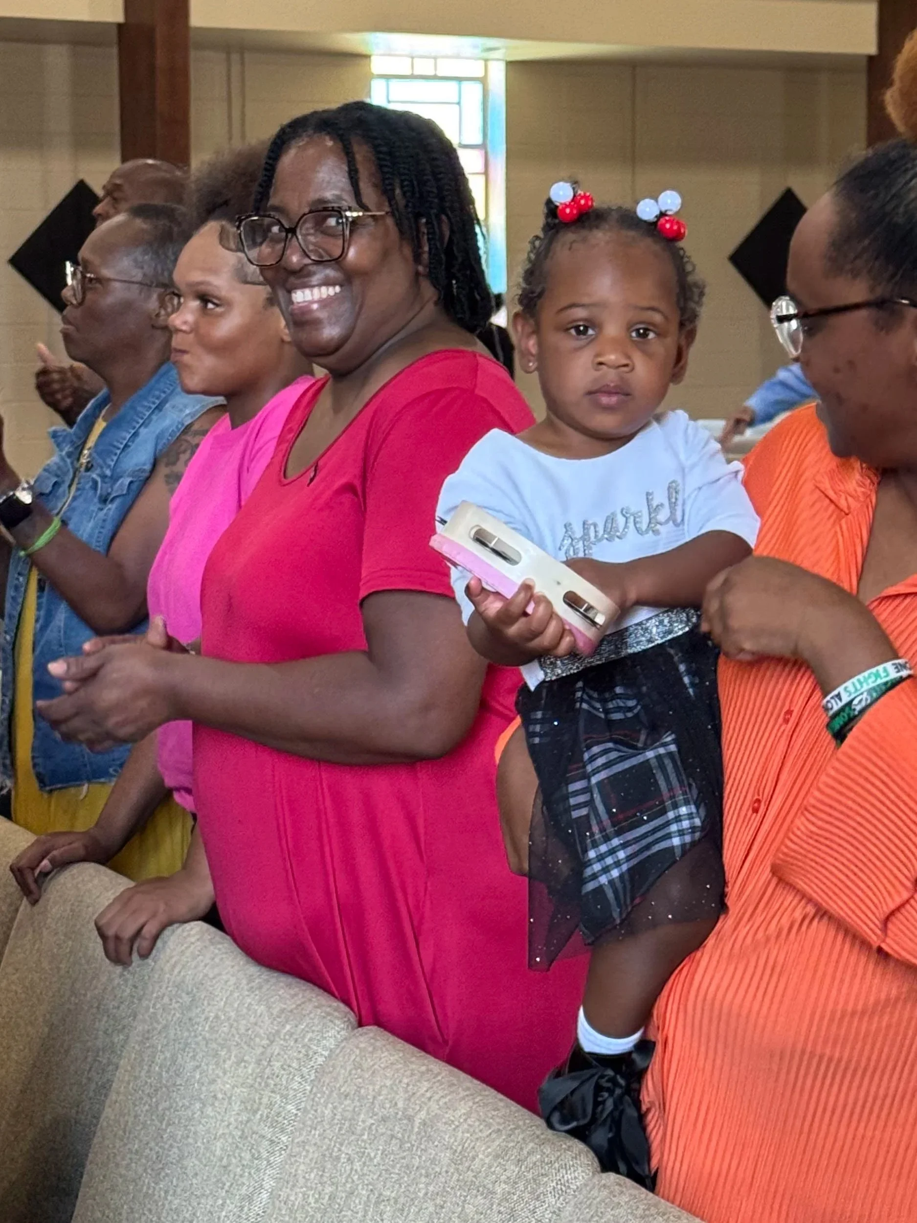 A group of diverse people standing in a church or similar setting, including a young girl holding a smartphone, a woman wearing glasses and a pink dress smiling, and other adults next to them.