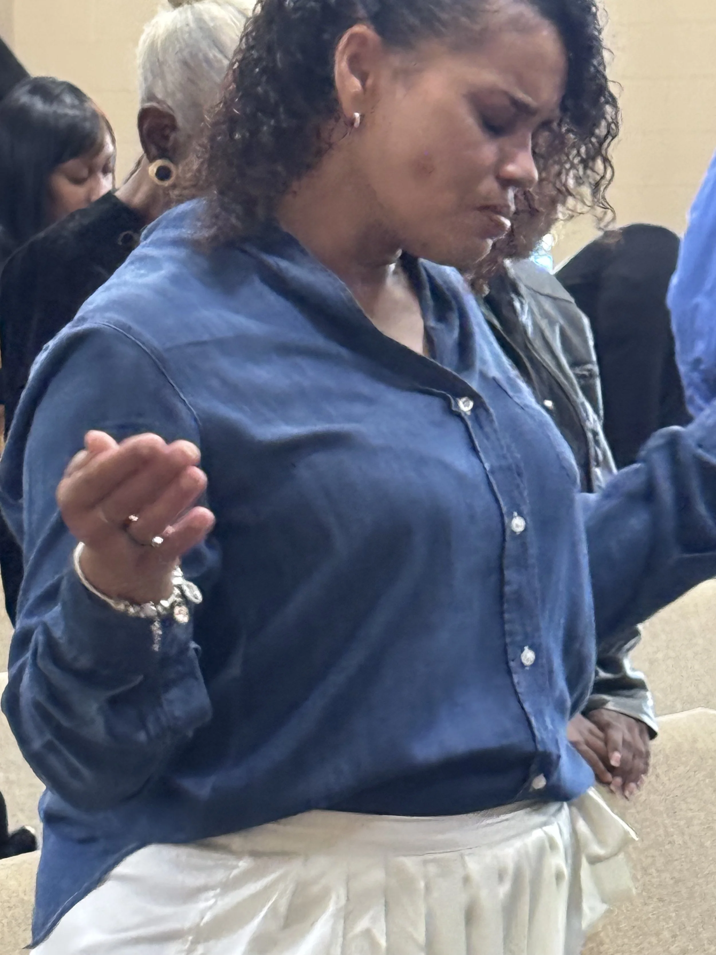 A woman with curly hair, wearing a blue denim shirt and white pants, is standing with her eyes closed and hands raised in a prayerful or meditative posture, surrounded by other people in a group setting.