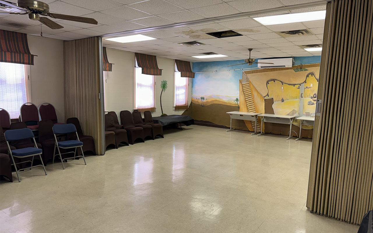 Large room with chairs aligned along the wall, windows with striped curtains, a mural of a desert landscape with palm trees, and tables against the wall. The ceiling has ceiling fans and some missing ceiling tiles.