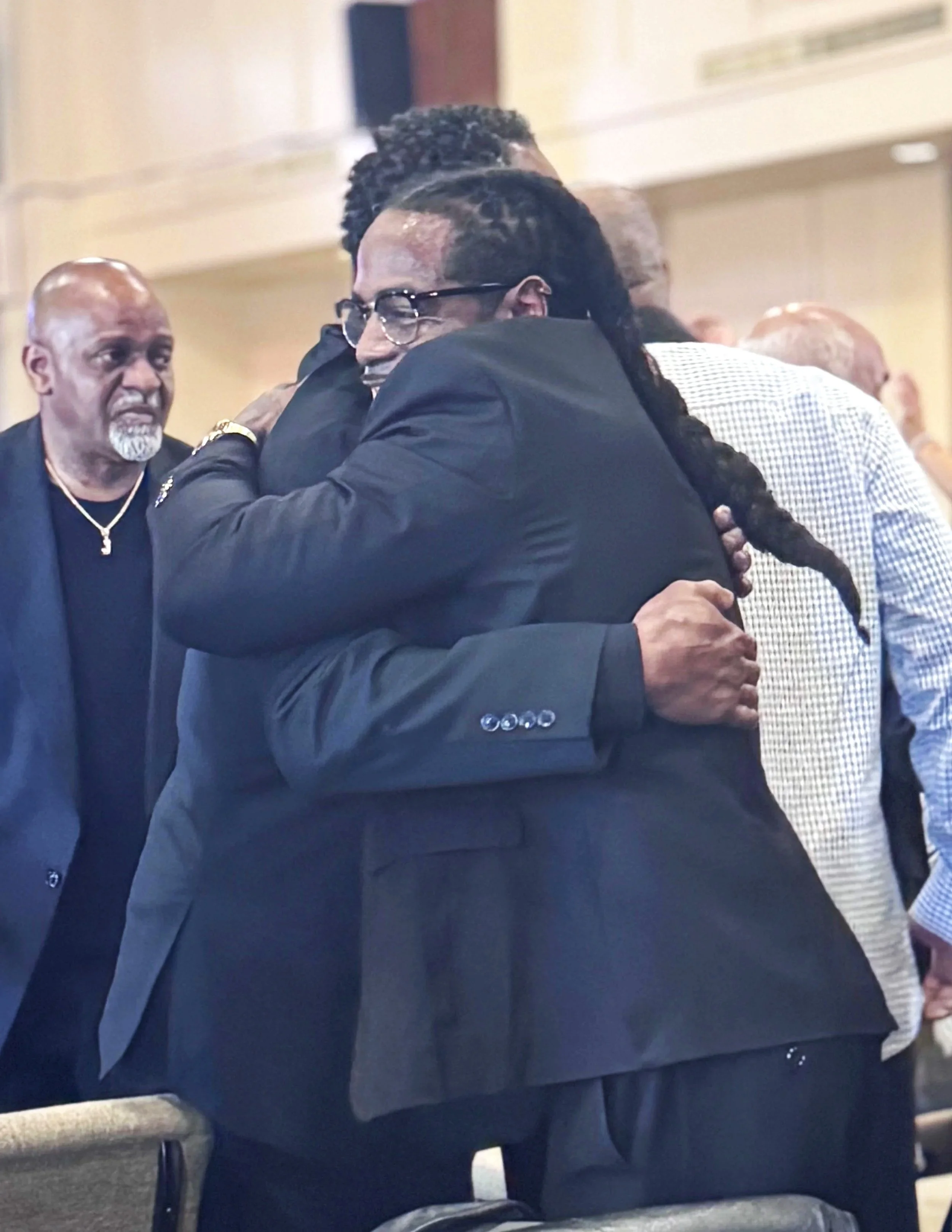 Two people hugging each other warmly at a gathering indoors, with others in the background.