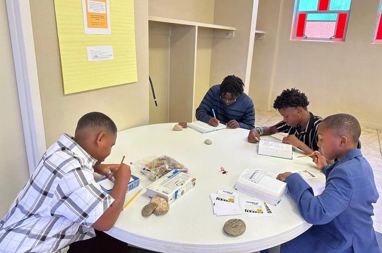 Five children sit around a white round table, reading books and writing with pencils in an indoor setting with beige walls and a window with red frames.
