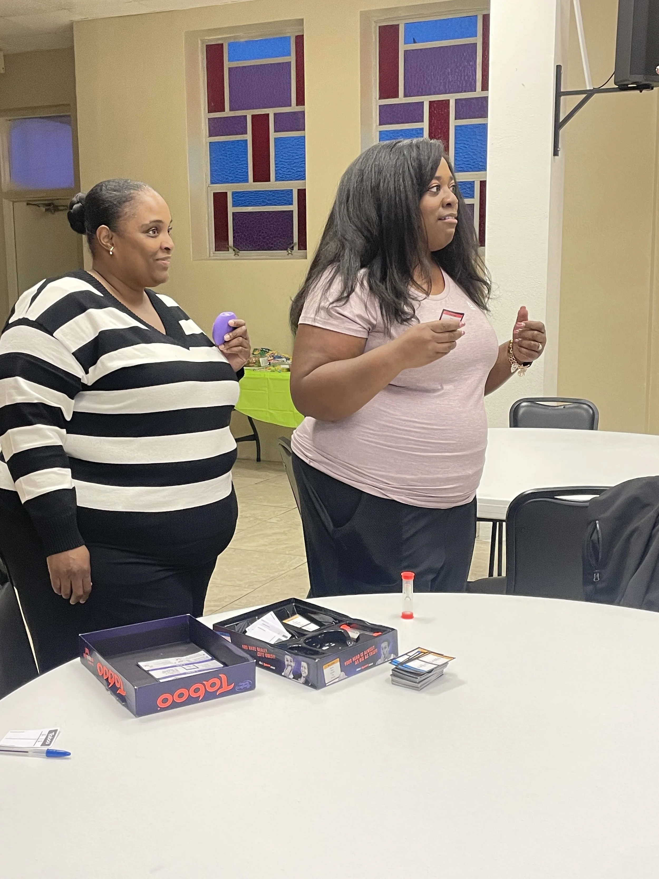 Two women standing by a table with board games and cards, inside a room with stained glass windows.