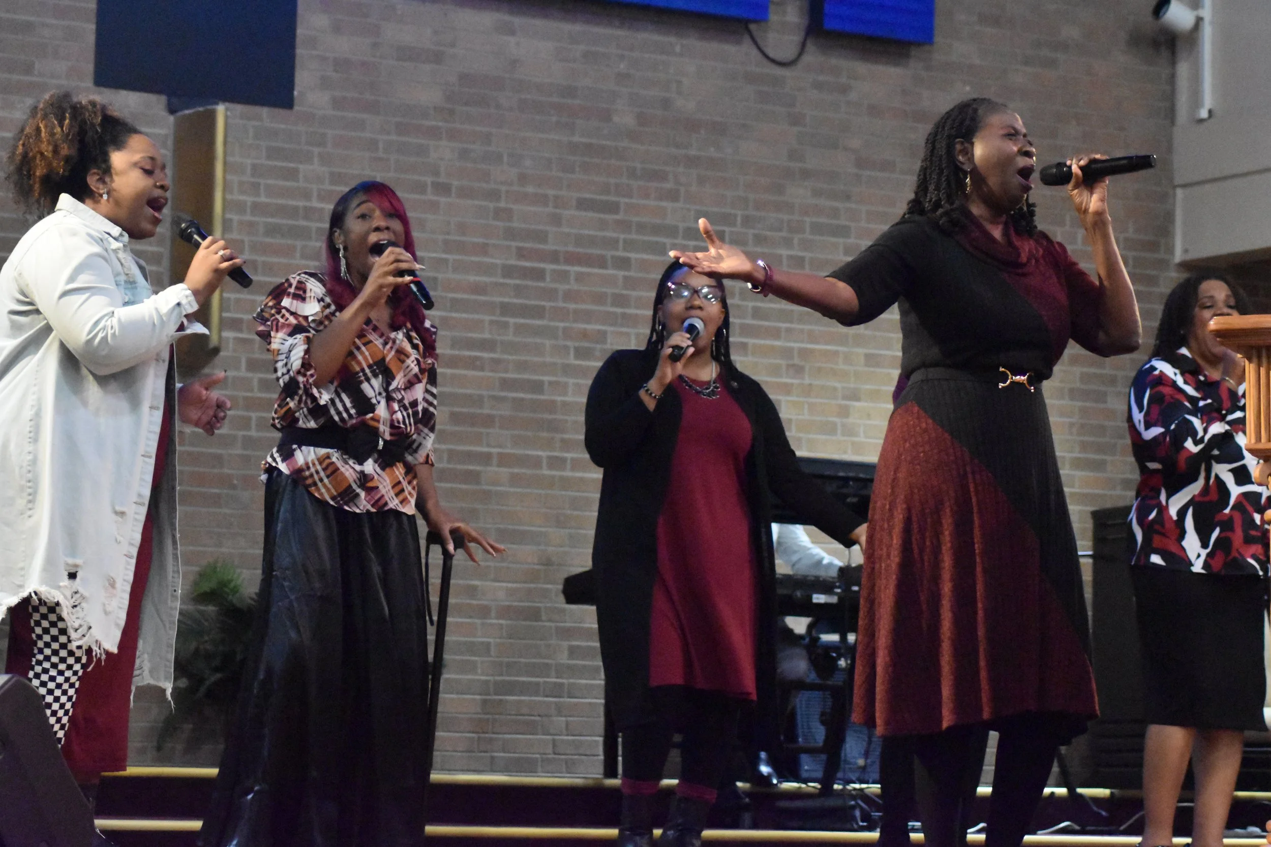 A group of women singing passionately on stage with microphones, with a brick wall background and a keyboard in the background.