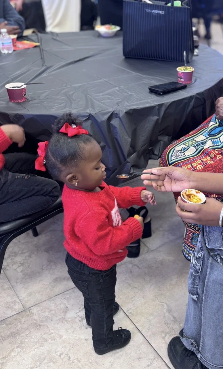 A young girl with a red bow in her hair and a red sweater is being fed food from a small container by an adult using a spoon, at a dining table covered with a black tablecloth in a social gathering.