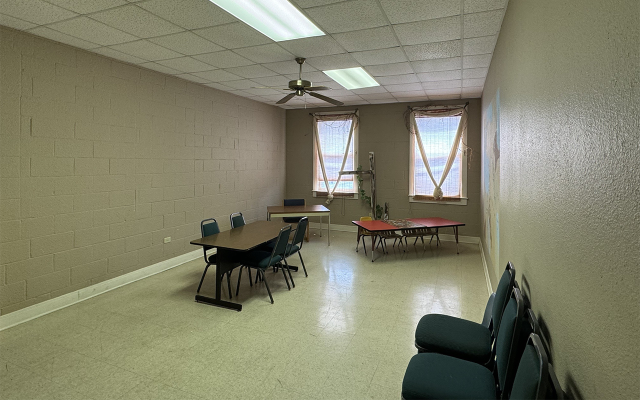 Classroom with three tables, chairs, a ceiling fan, and two windows with curtains.