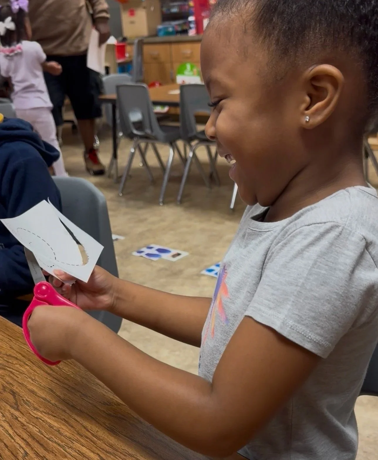 A young girl smiling and cutting a piece of paper with pink scissors in a classroom or craft room setting.