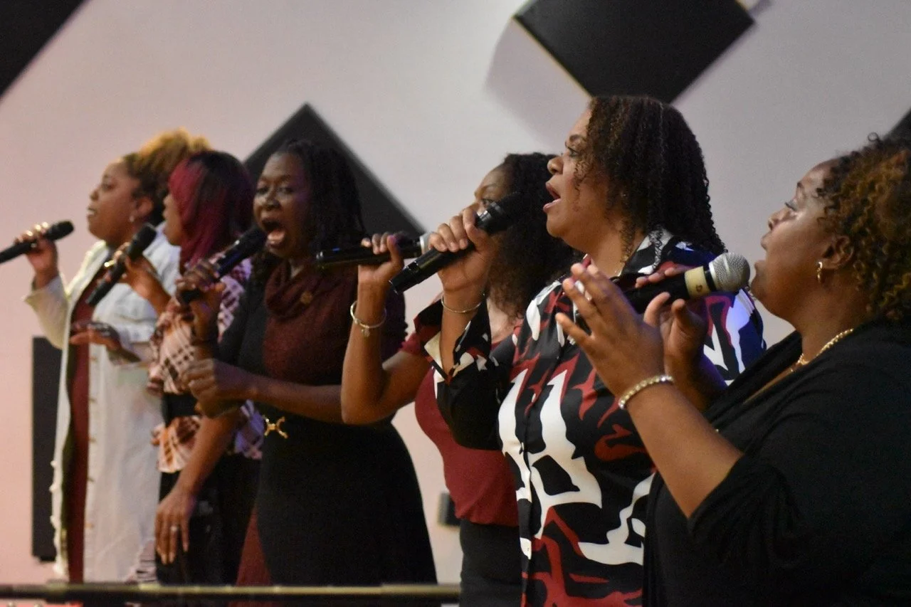 A group of women singing into microphones on stage during a performance or rehearsal.