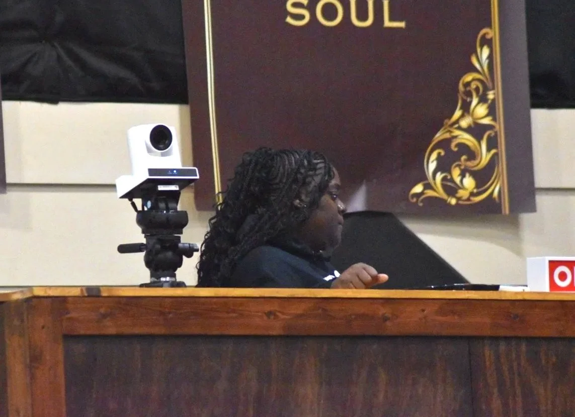 A woman with long black braided hair working at a desk with a camera on a tripod next to her. There is a decorative sign behind her with the word 'SOUL' on it.