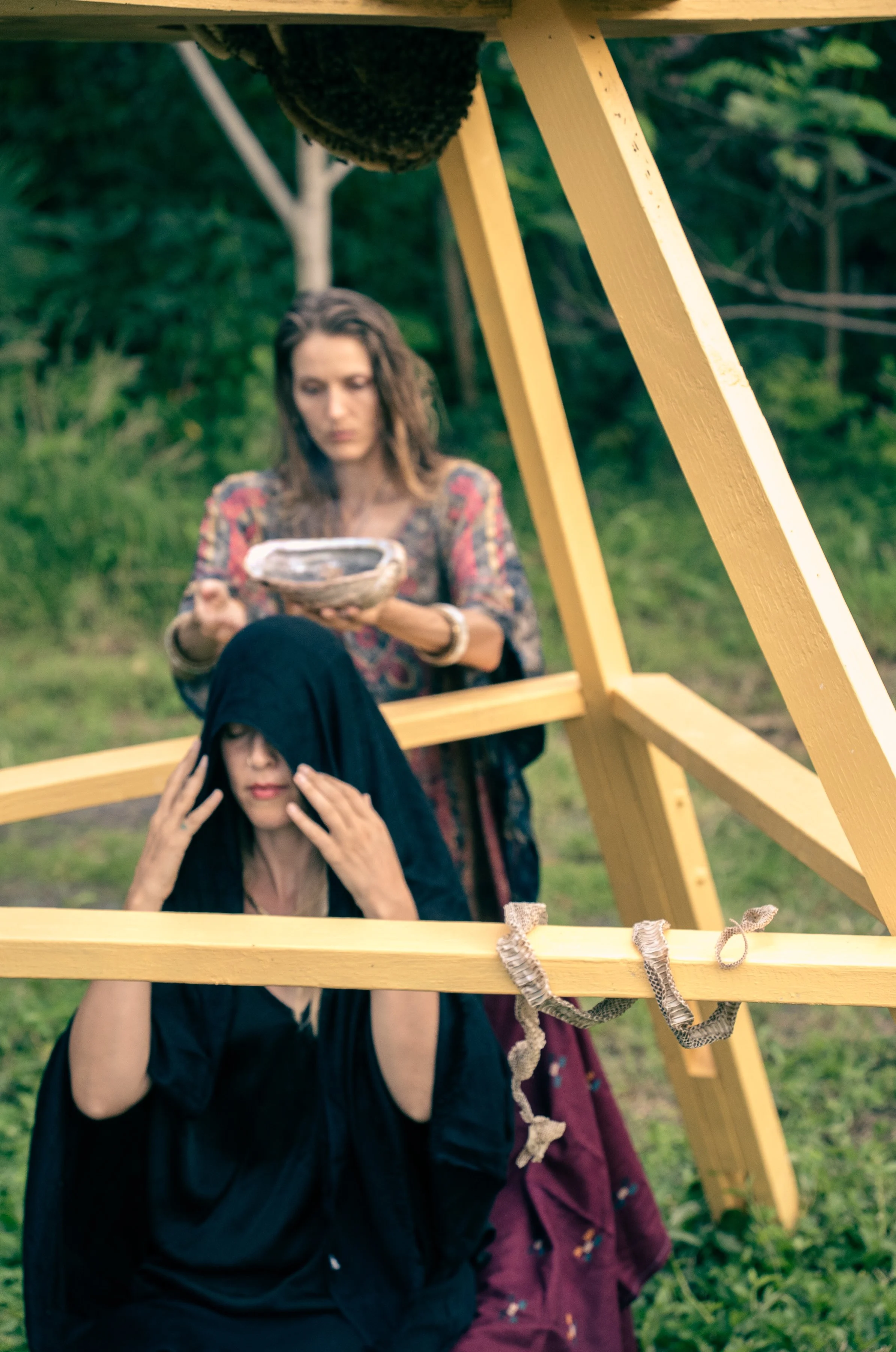 Two women outdoors with a yellow wooden structure. One woman is sitting and covering her face with a black garment, and the other woman standing behind her is holding a small bowl and looking down.