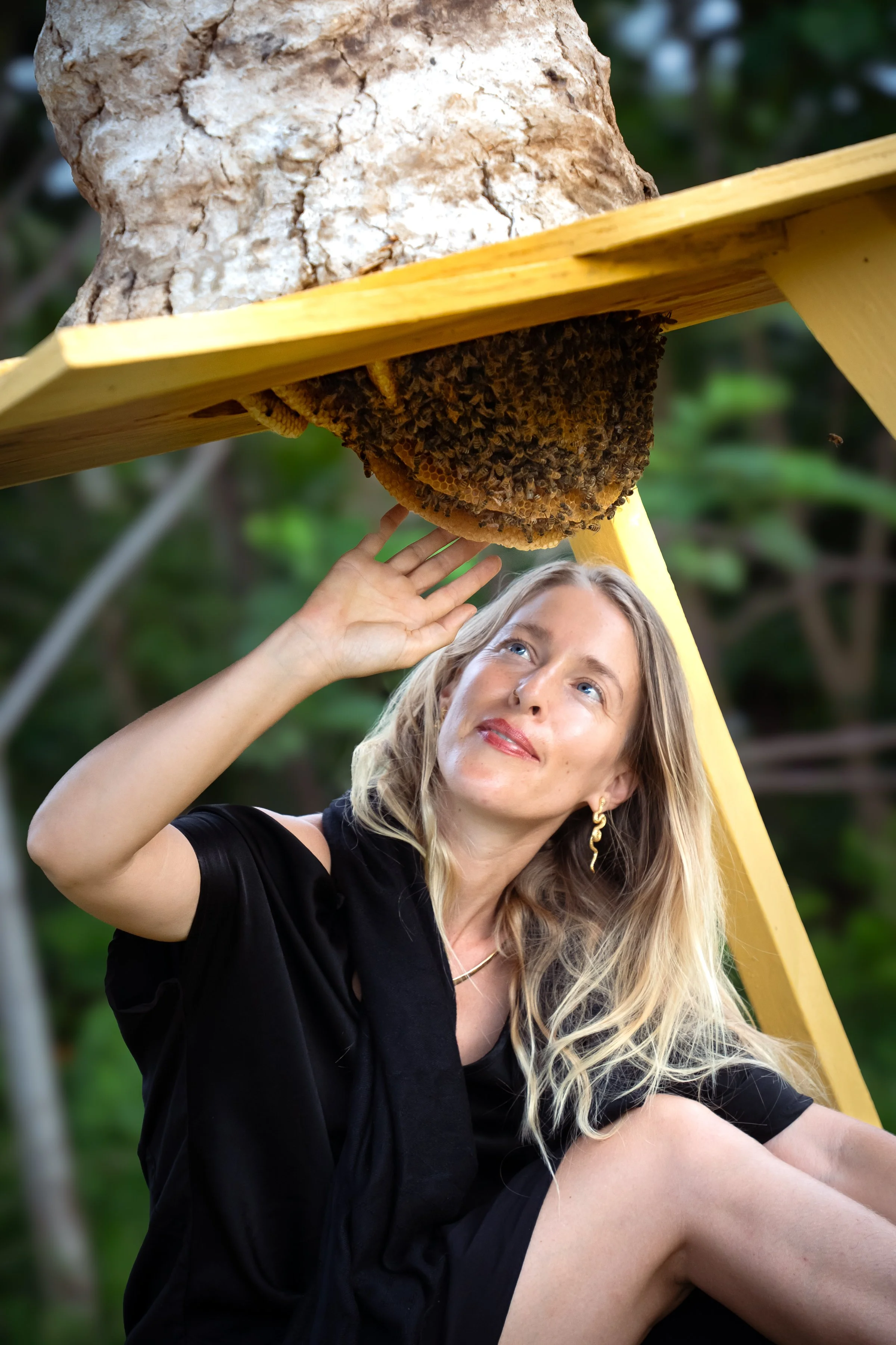 A woman with blonde hair, wearing a black dress and earrings, sitting outdoors. She is touching a beehive hanging from a yellow wooden structure, with a background of blurred green foliage.