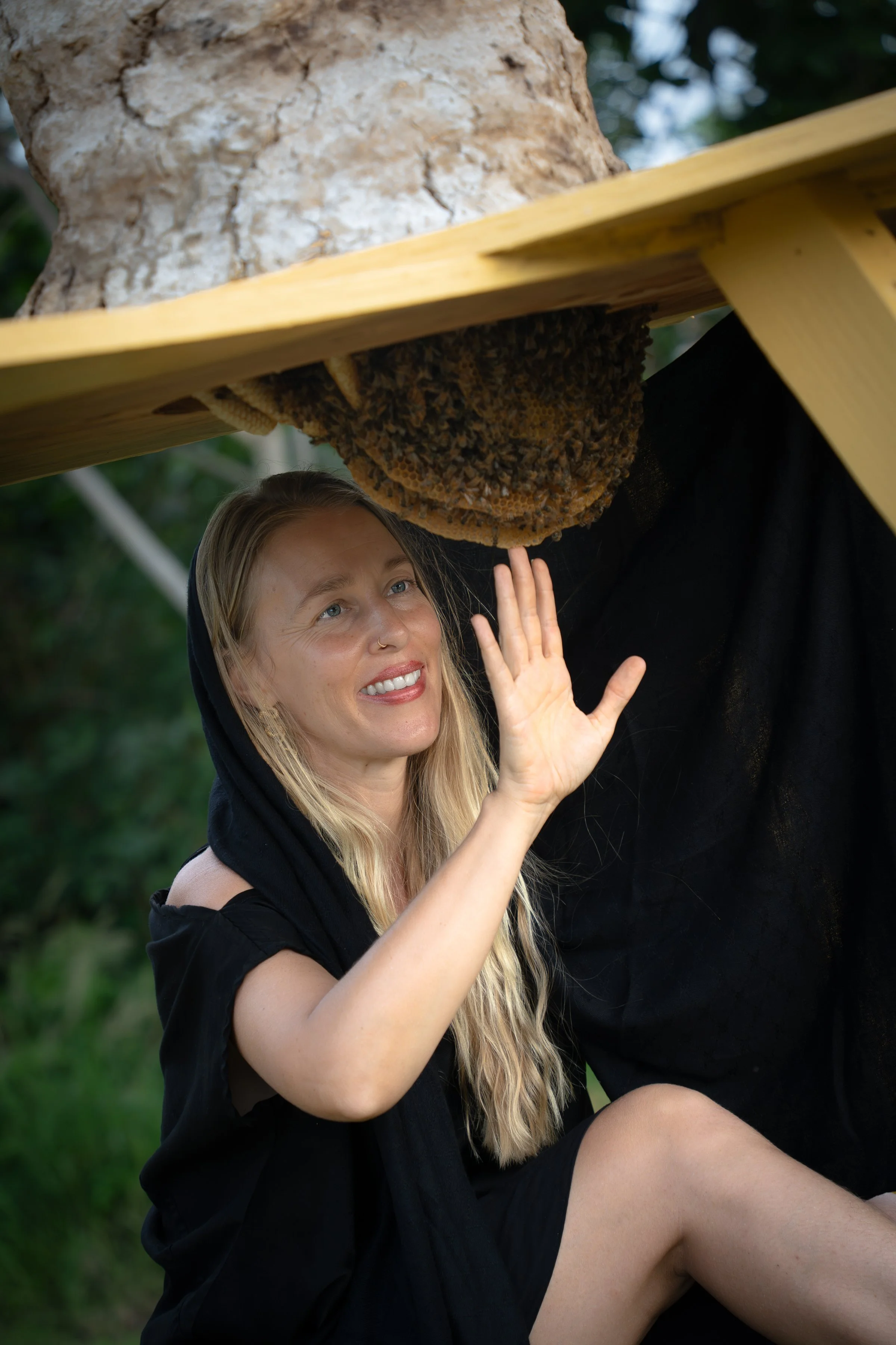 A woman with blonde hair and a black hooded garment, smiling and waving, sitting outdoors beneath a honey bee hive hanging from a wooden structure, with a background of greenery.