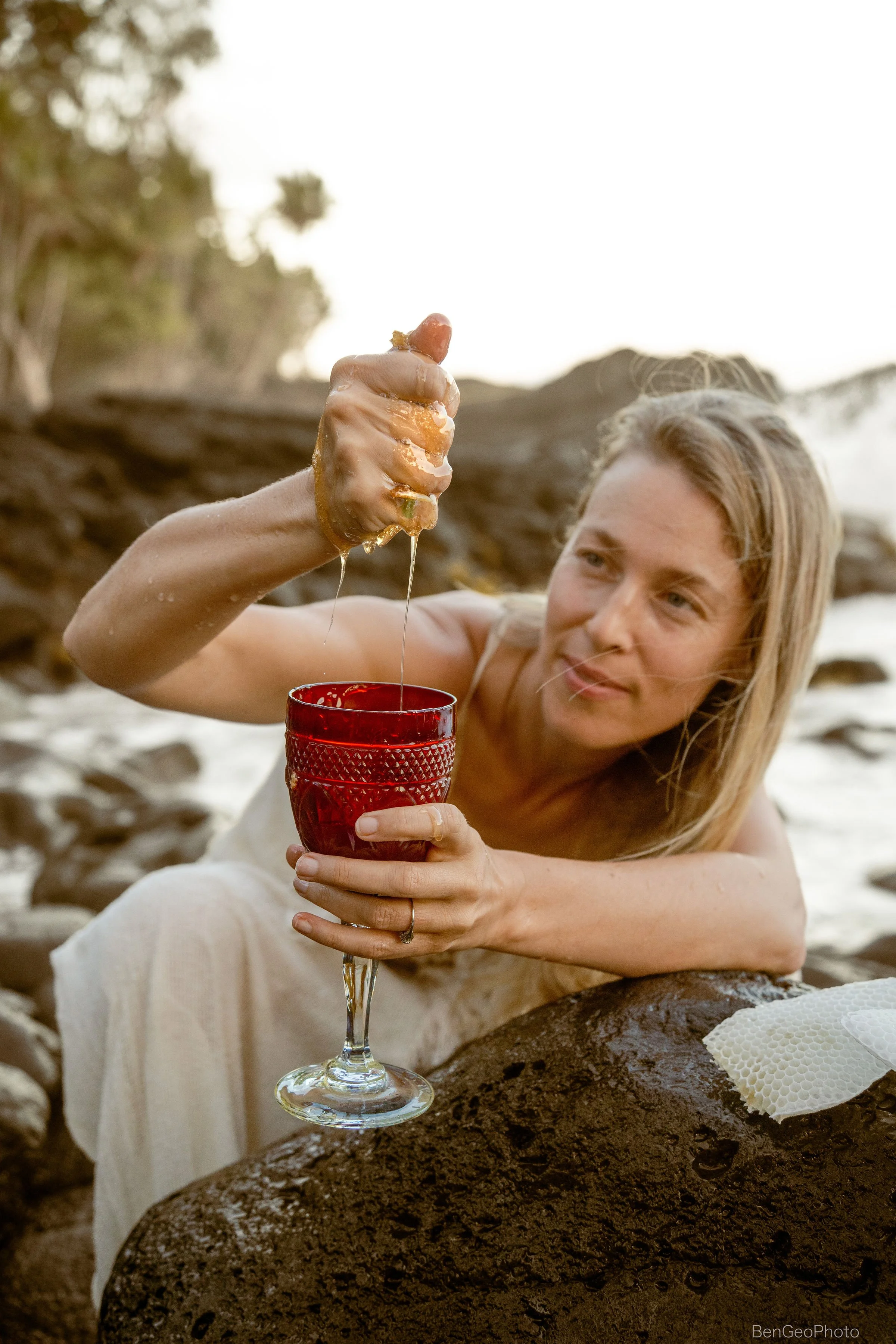 A woman with blonde hair lying on rocks at the beach, holding a glass of red wine, and pouring honey from her hand.