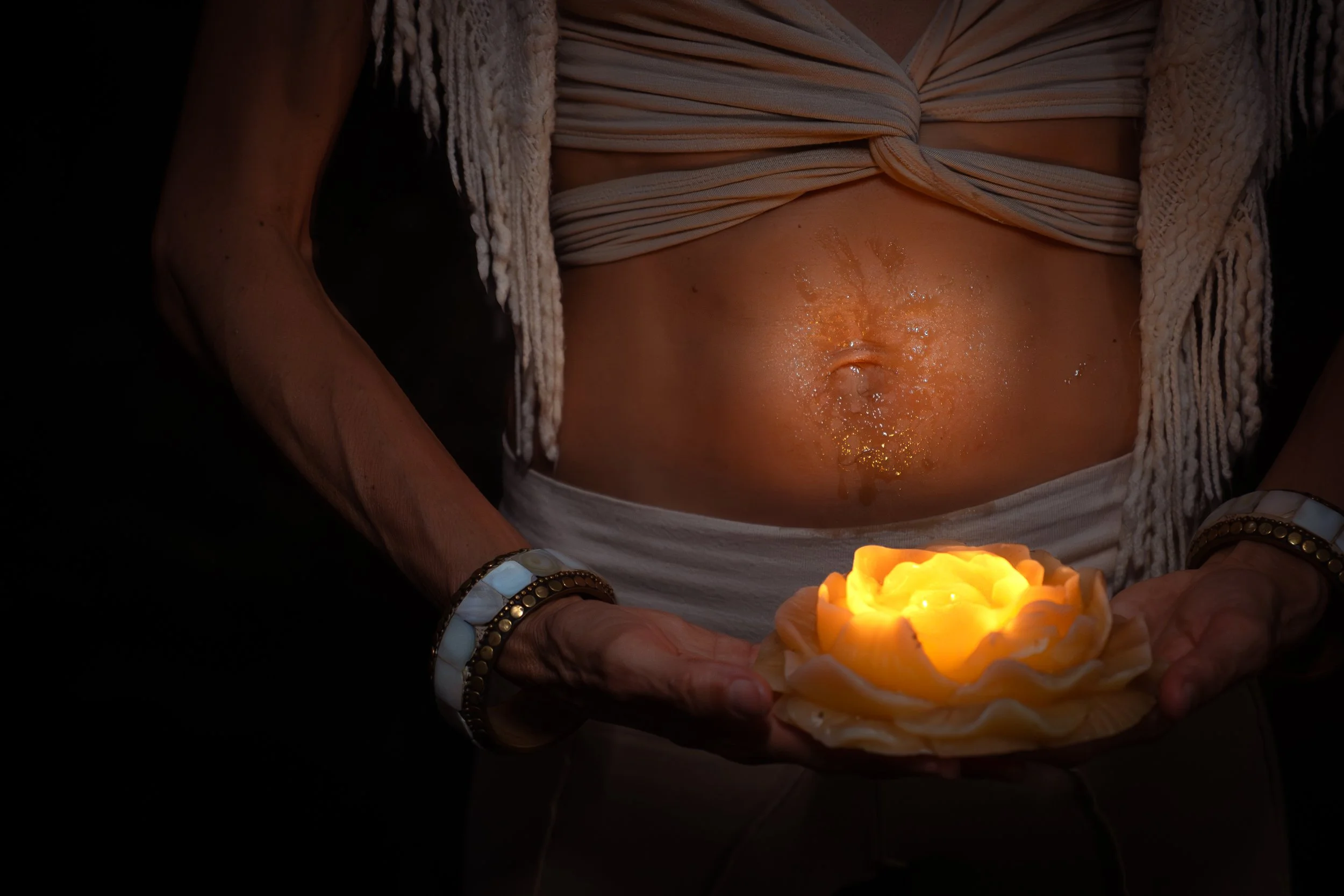 Close-up of a woman’s bare abdomen, holding a lit yellow candle in a floral-shaped holder. She is wearing a striped top tied in the front and a knitted cardigan. The setting appears to be dimly lit.