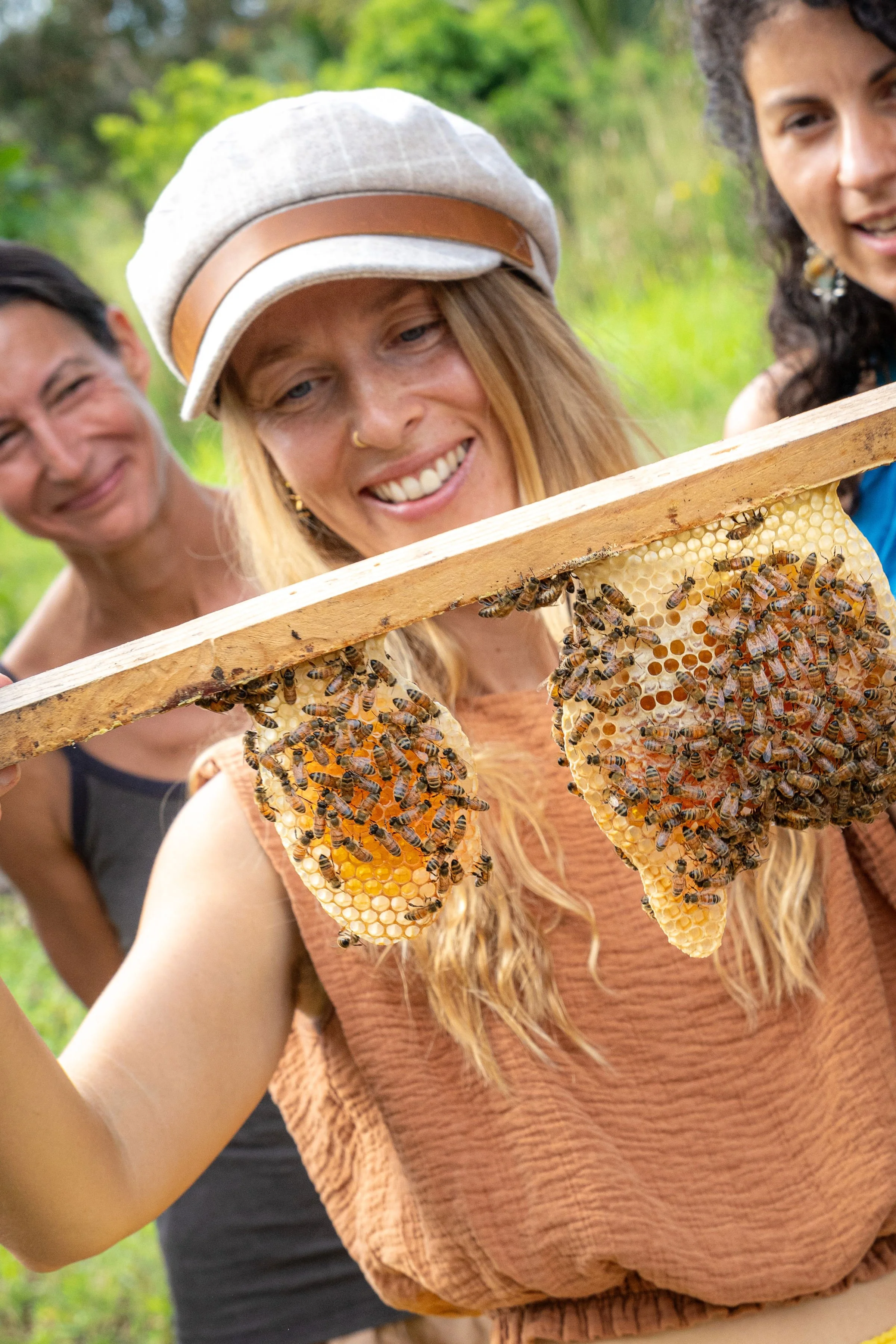A group of people examining a honeycomb frame with bees on it outdoors, with green foliage in the background.