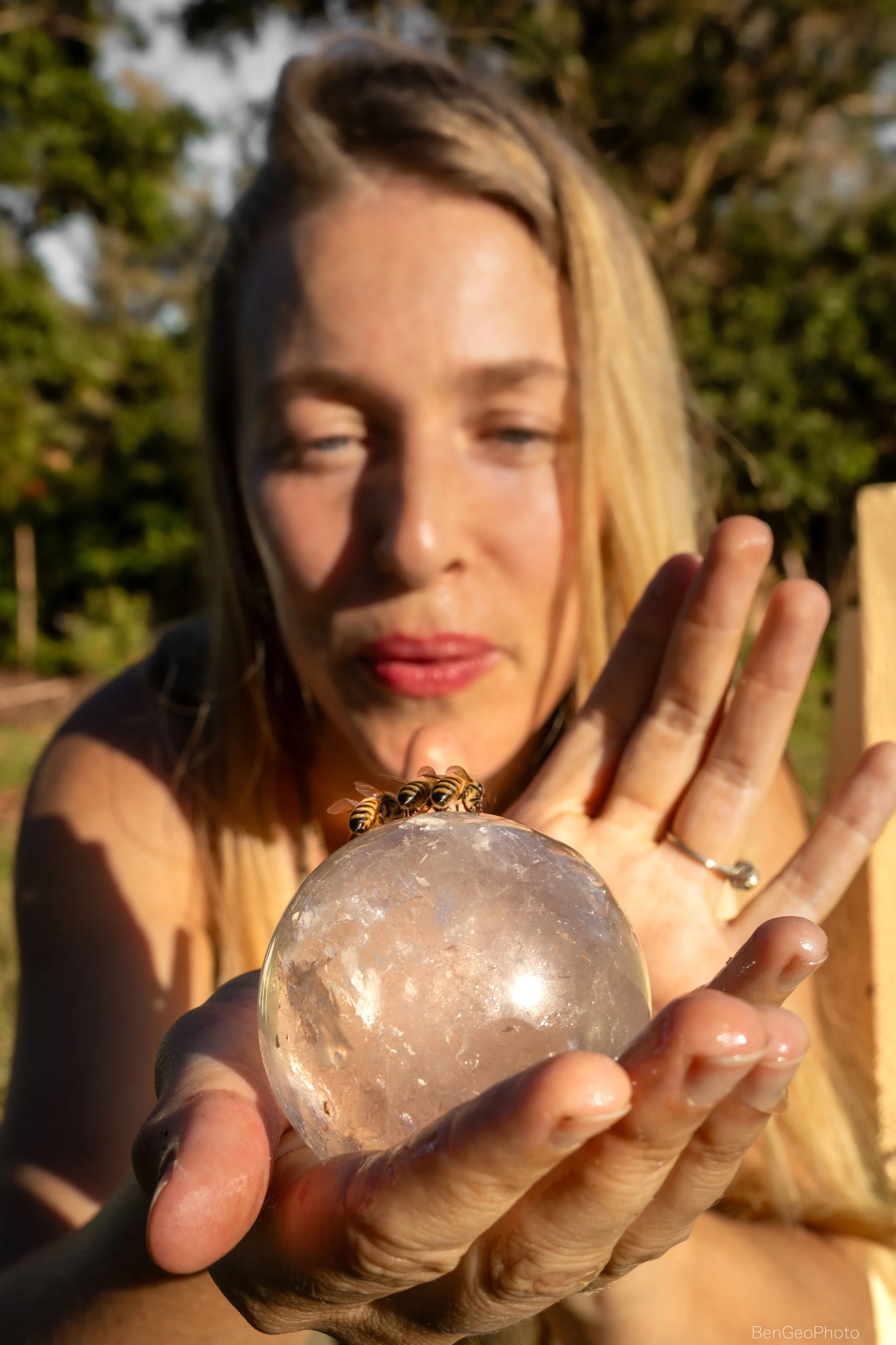 A woman with blonde hair holding a clear crystal ball with a bee on top, outdoors with trees in the background, during daylight.