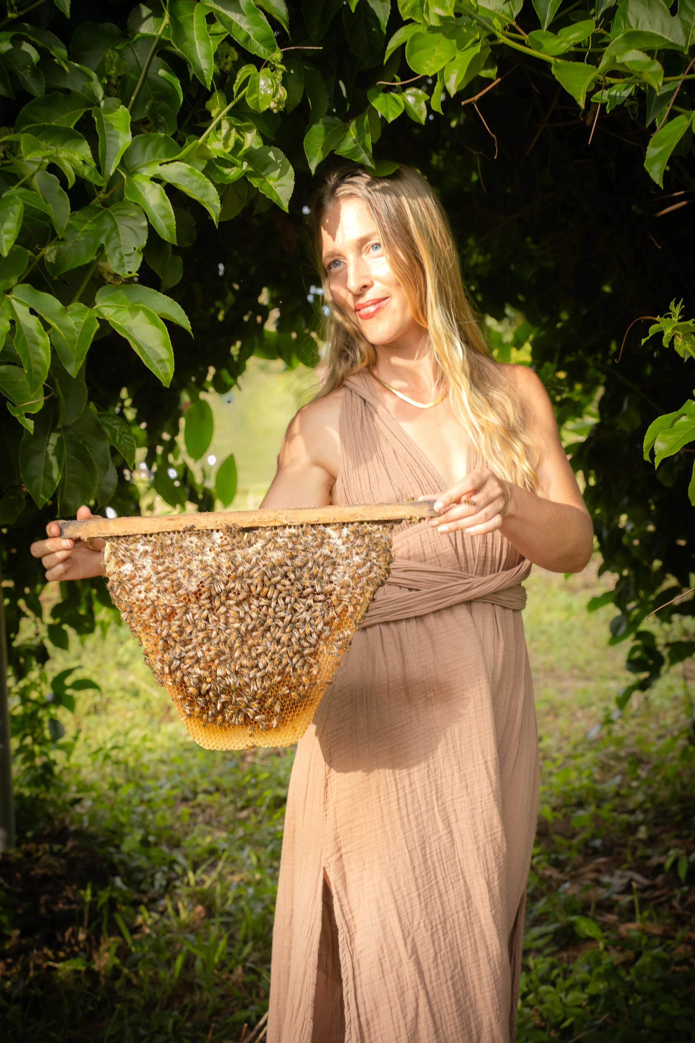 A woman with blonde hair in a beige dress holding a honeycomb frame, standing in a lush green hive environment.