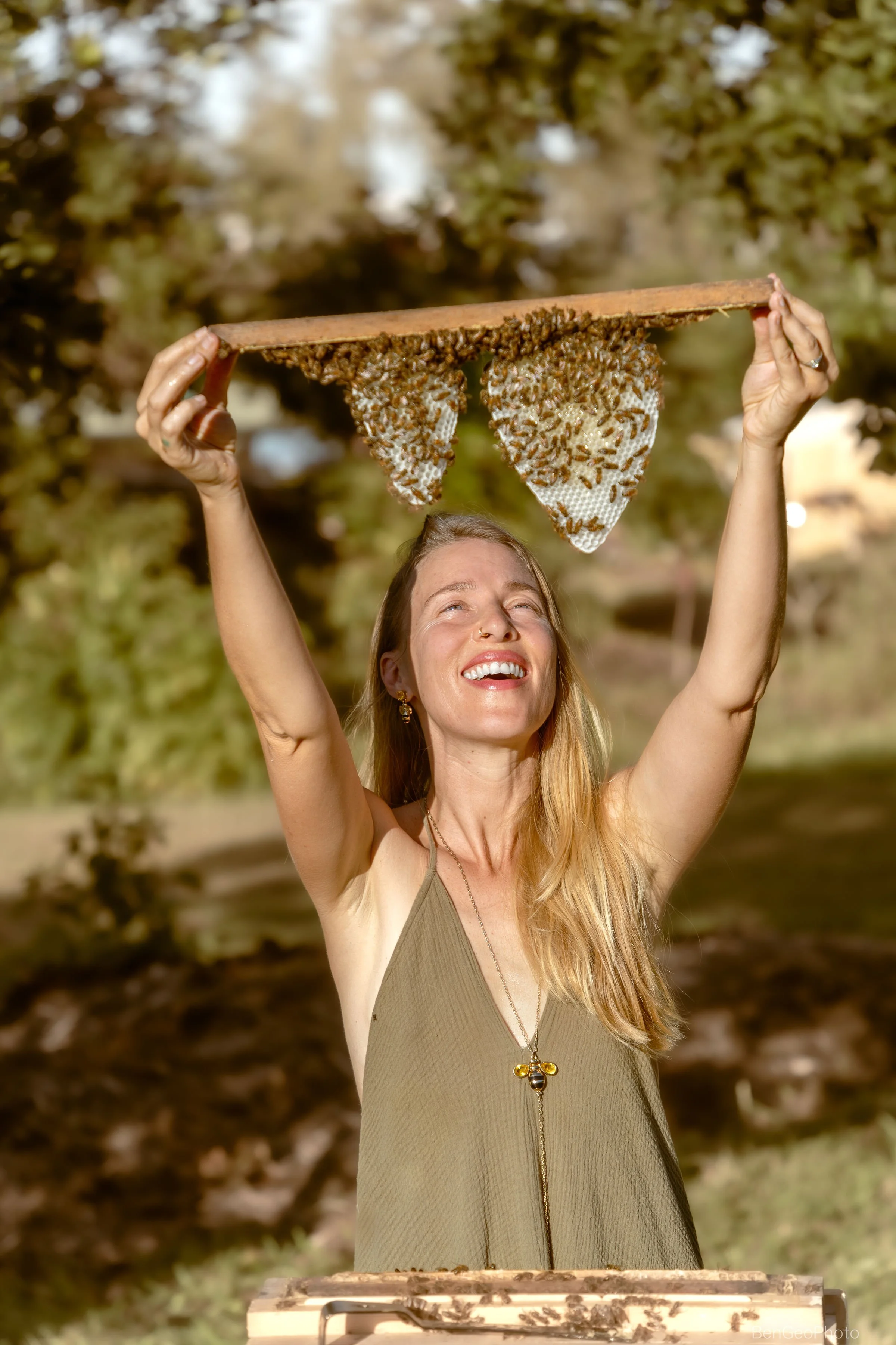 A woman with long blond hair, smiling, holds a framed honeycomb with bees above her head outdoors, with trees and dappled sunlight in the background.