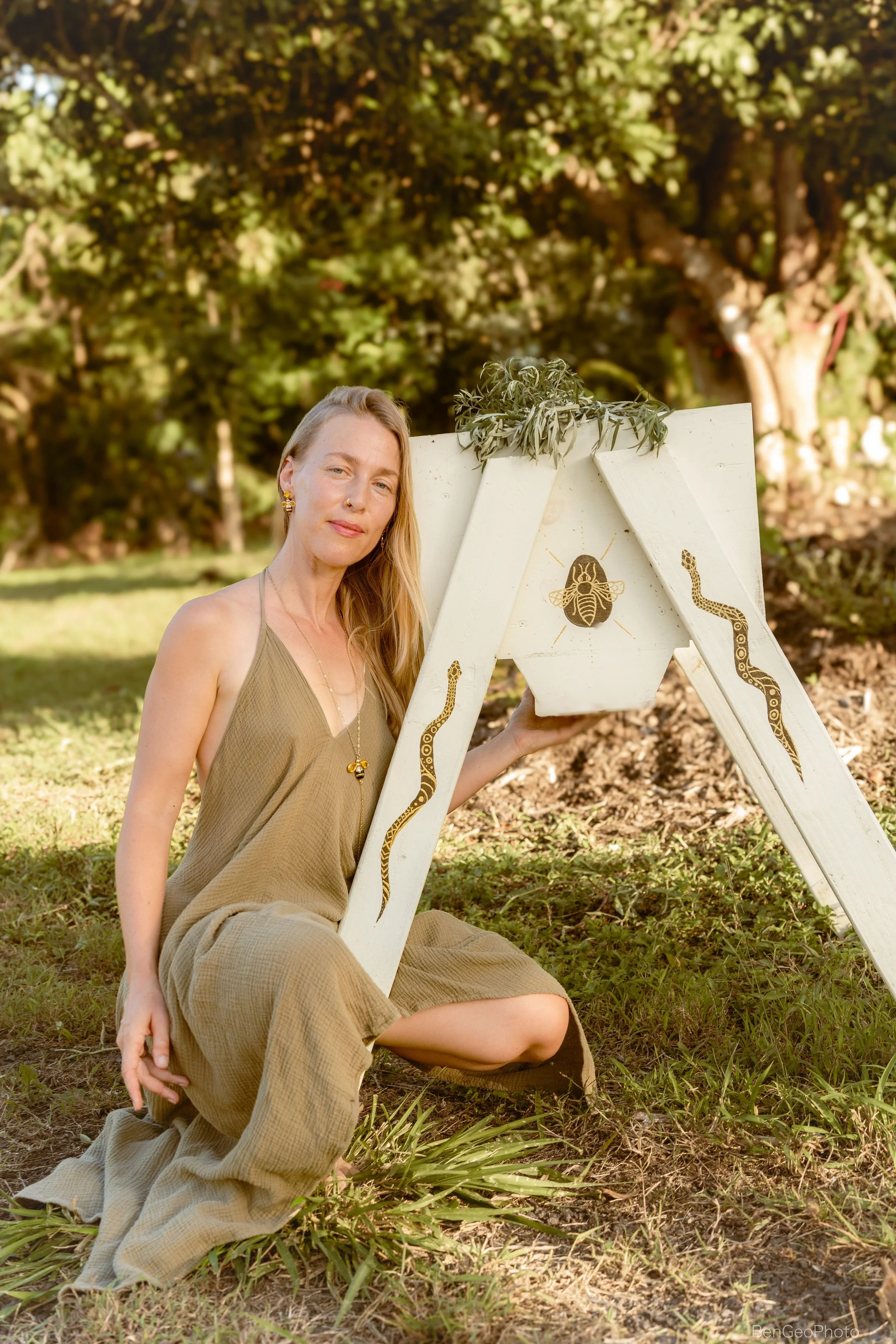 A woman with long blonde hair, wearing a beige outfit and gold jewelry, sitting on the grass outdoors, leaning against a decorative wooden easel with snake and bee art, surrounded by greenery and trees.
