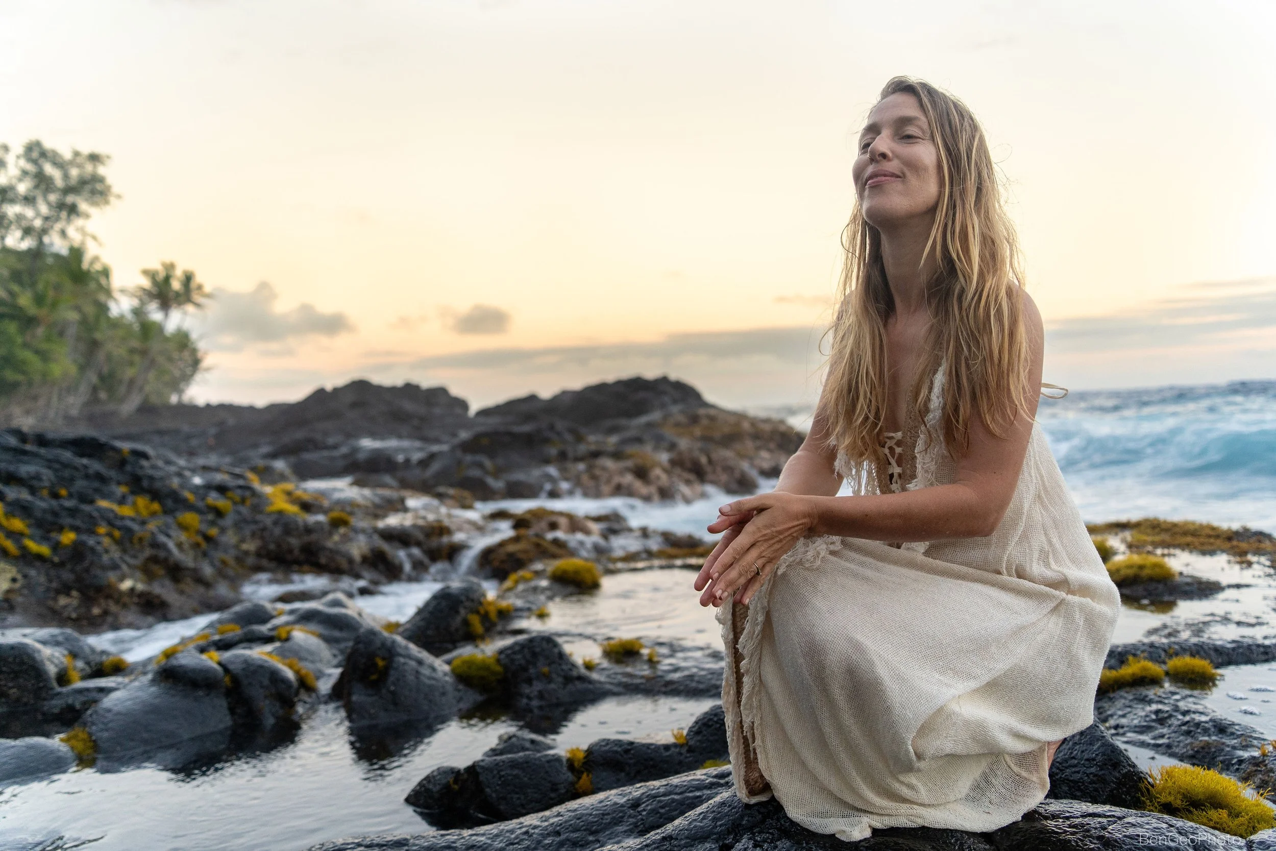 A woman with long blonde hair sitting on rocks by the beach on the Big Island Hawaii during sunset, wearing a light-colored dress, with her eyes closed and a peaceful expression.