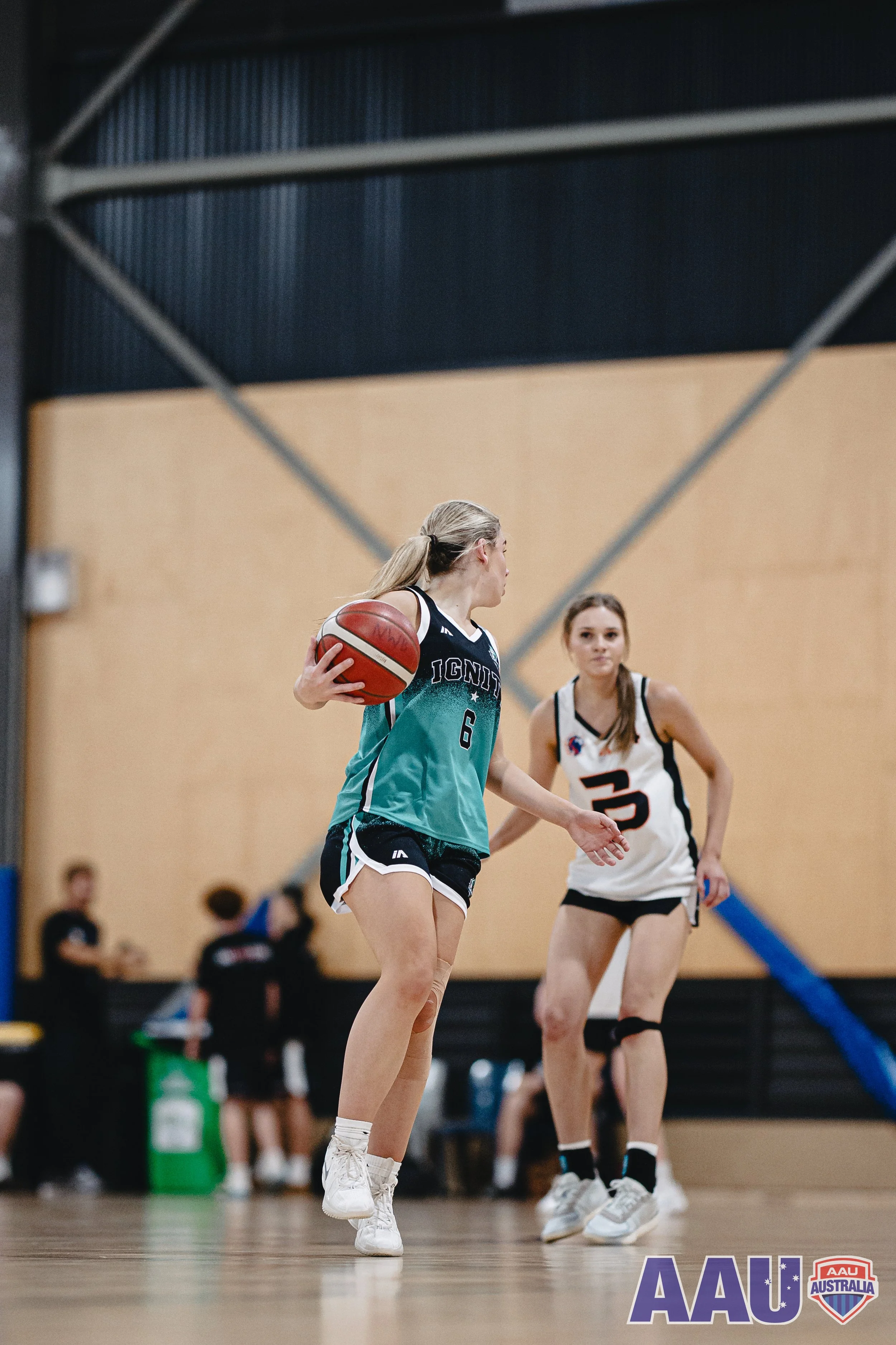 Girls playing basketball in an indoor gym, one holding a basketball, wearing teal and black uniform, and another in a white uniform with black trim, with the AAU Australia logo in the bottom right corner.