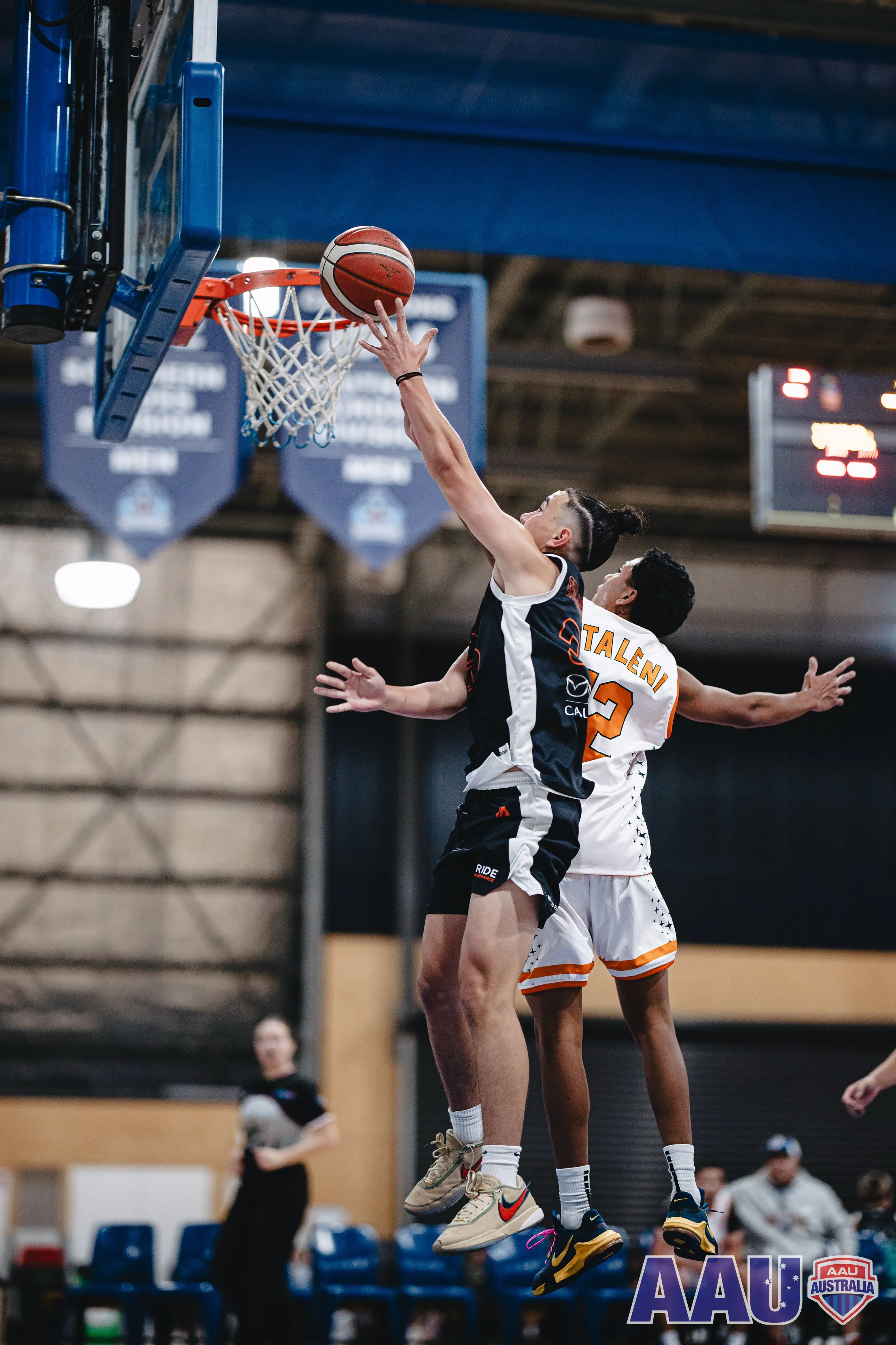 Two basketball players are jumping to score or block a shot during a game. One player in black is reaching towards the basket, while the other in white with orange accents is trying to defend. The game is played indoors, with a scoreboard and spectators in the background.