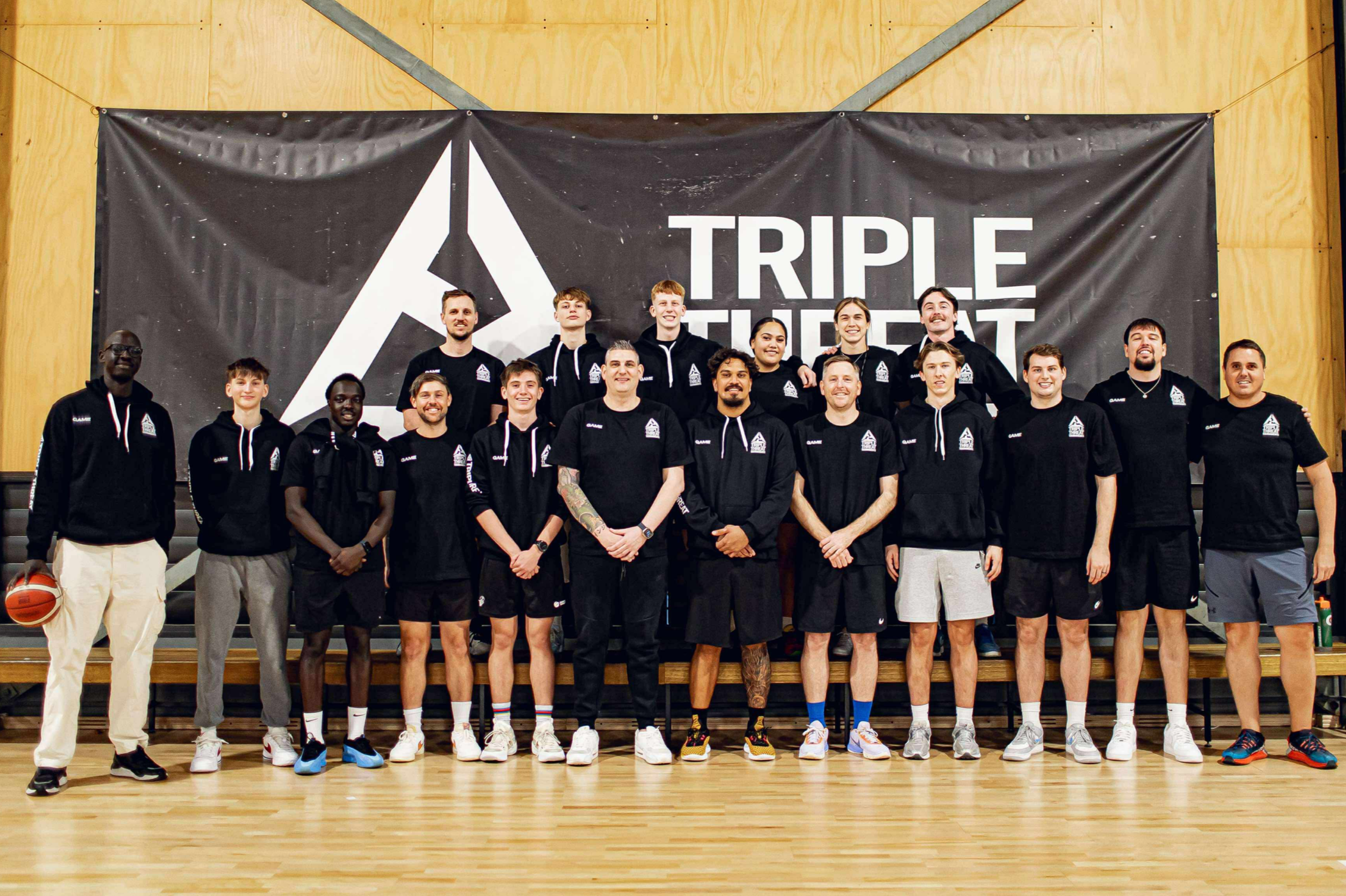 A group of basketball players and coaches posing for a team photo in front of a black banner with a large logo and the words 'TRIPLE THREAT'. They are inside a gymnasium with a wooden floor and walls.