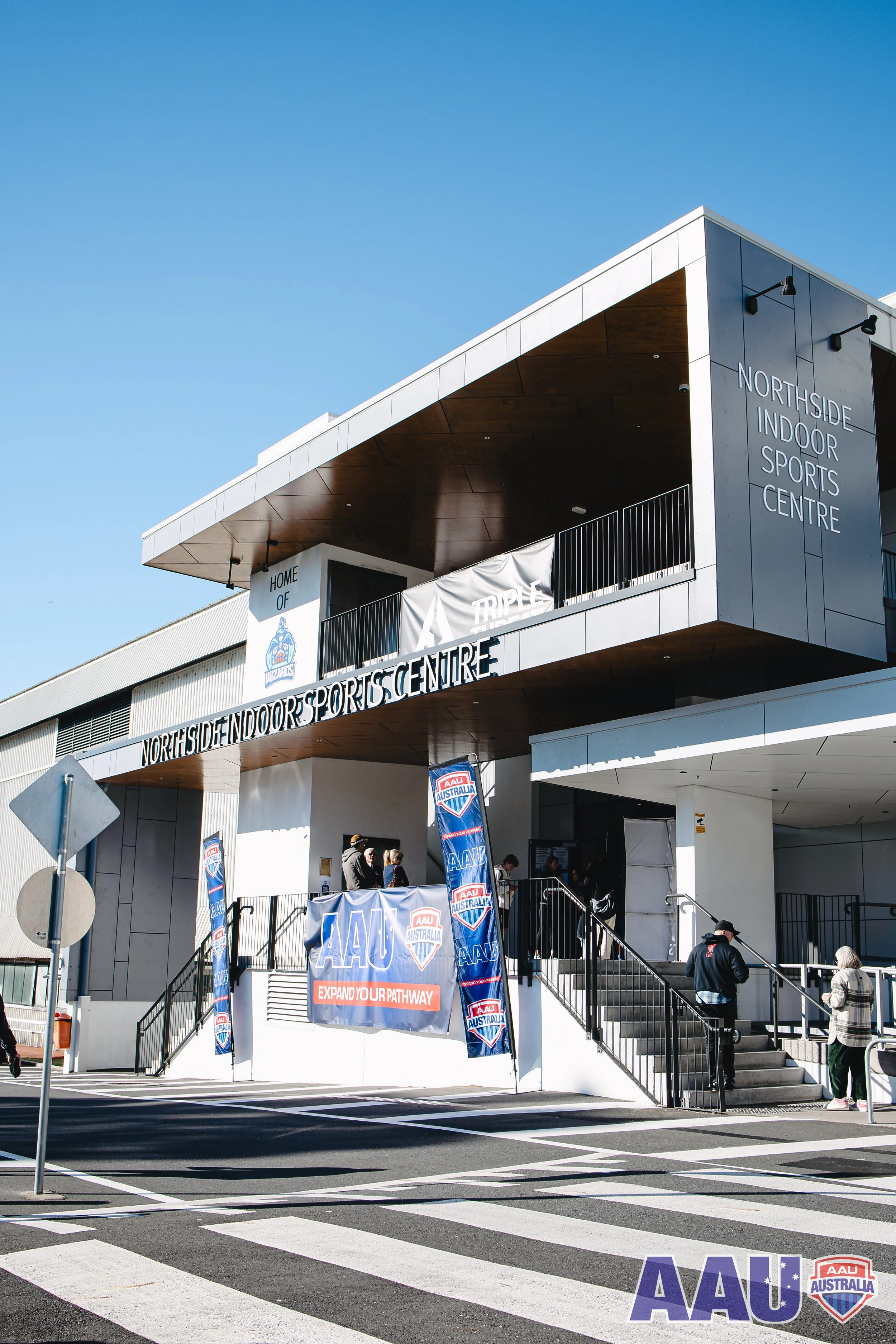 Exterior of the Northside Indoor Sports Centre with signage, banners, and several people walking in front of the entrance on a sunny day.