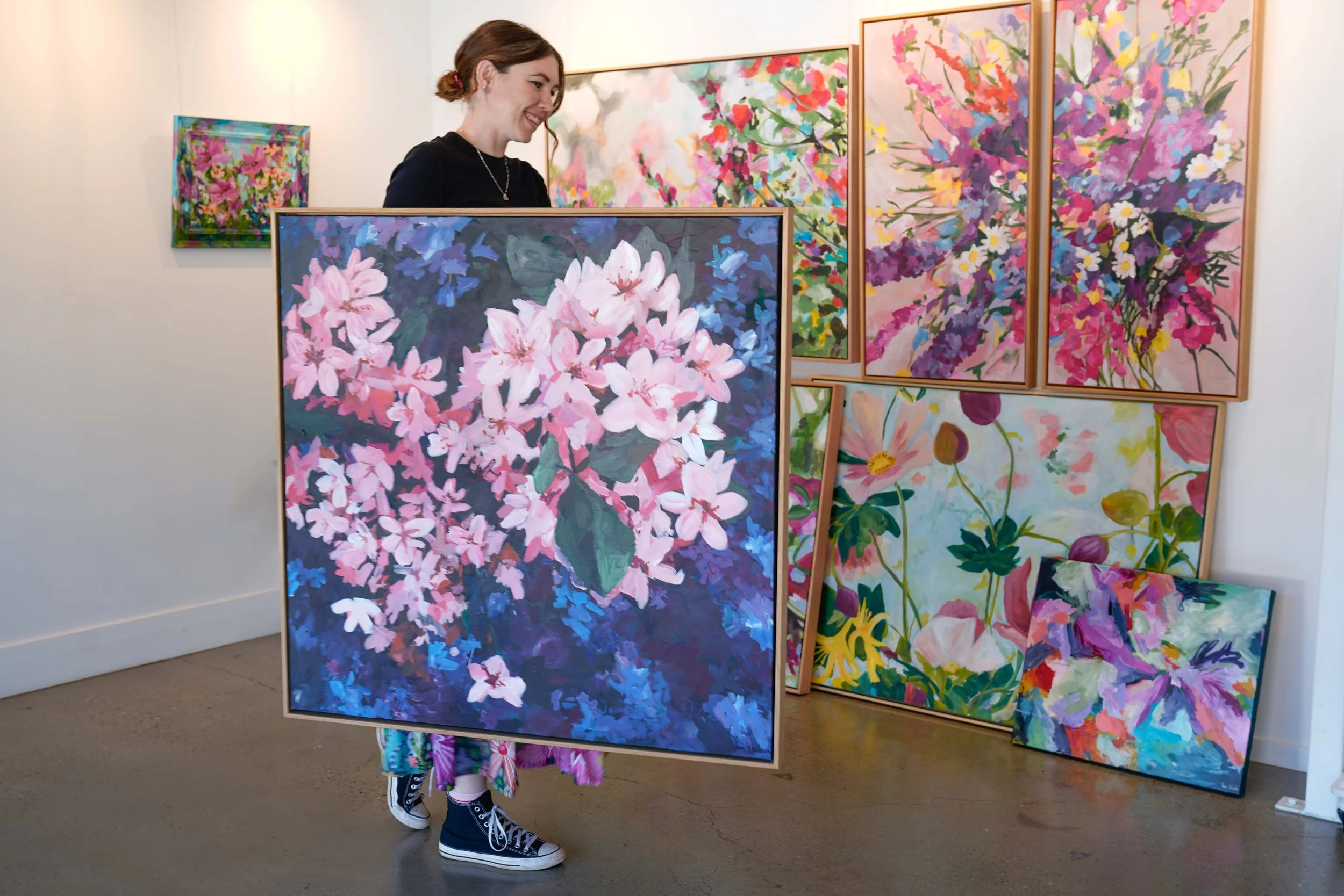 A woman with red hair, black shirt, and colorful patterned pants is carrying a large painting of pink flowers in an art gallery. There are other floral paintings on the wall behind her.