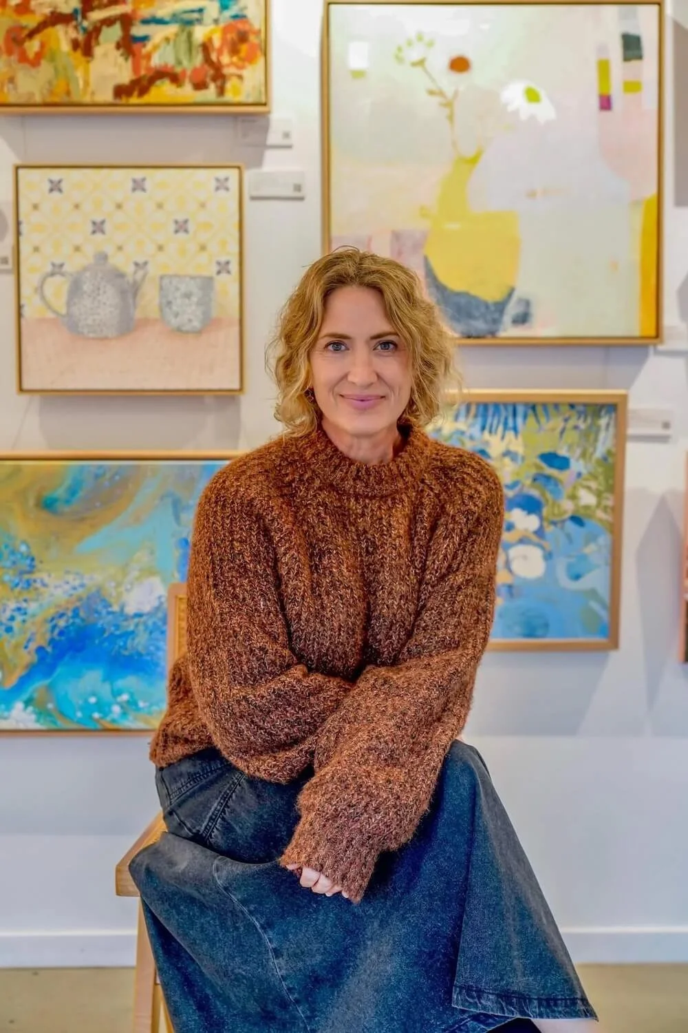 A woman with curly blonde hair sitting in an art gallery among colorful abstract paintings.