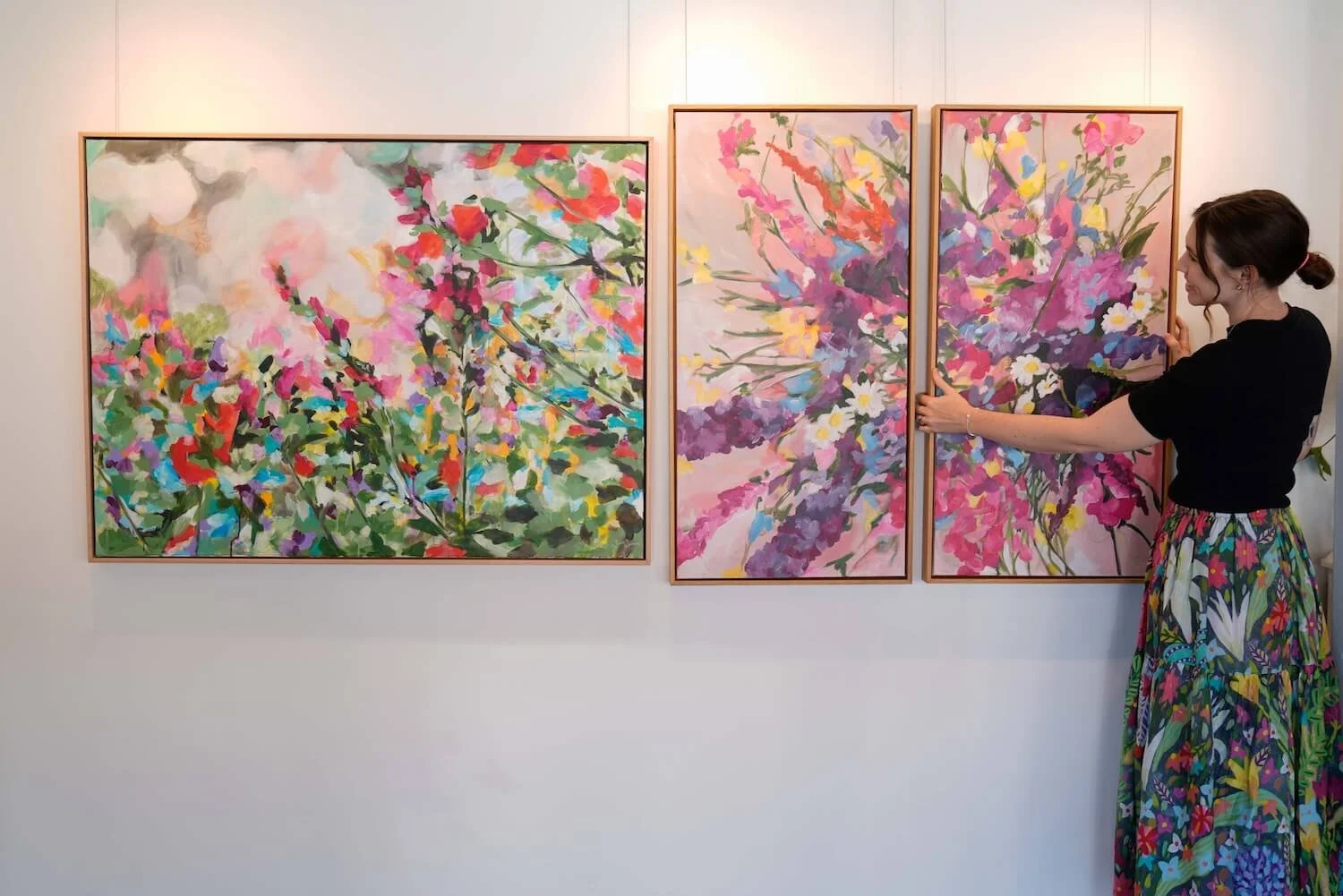 A woman in a black top and a long colorful floral skirt is arranging or examining a set of four abstract floral paintings with pink, purple, yellow, green, and white colors hanging on a white gallery wall.