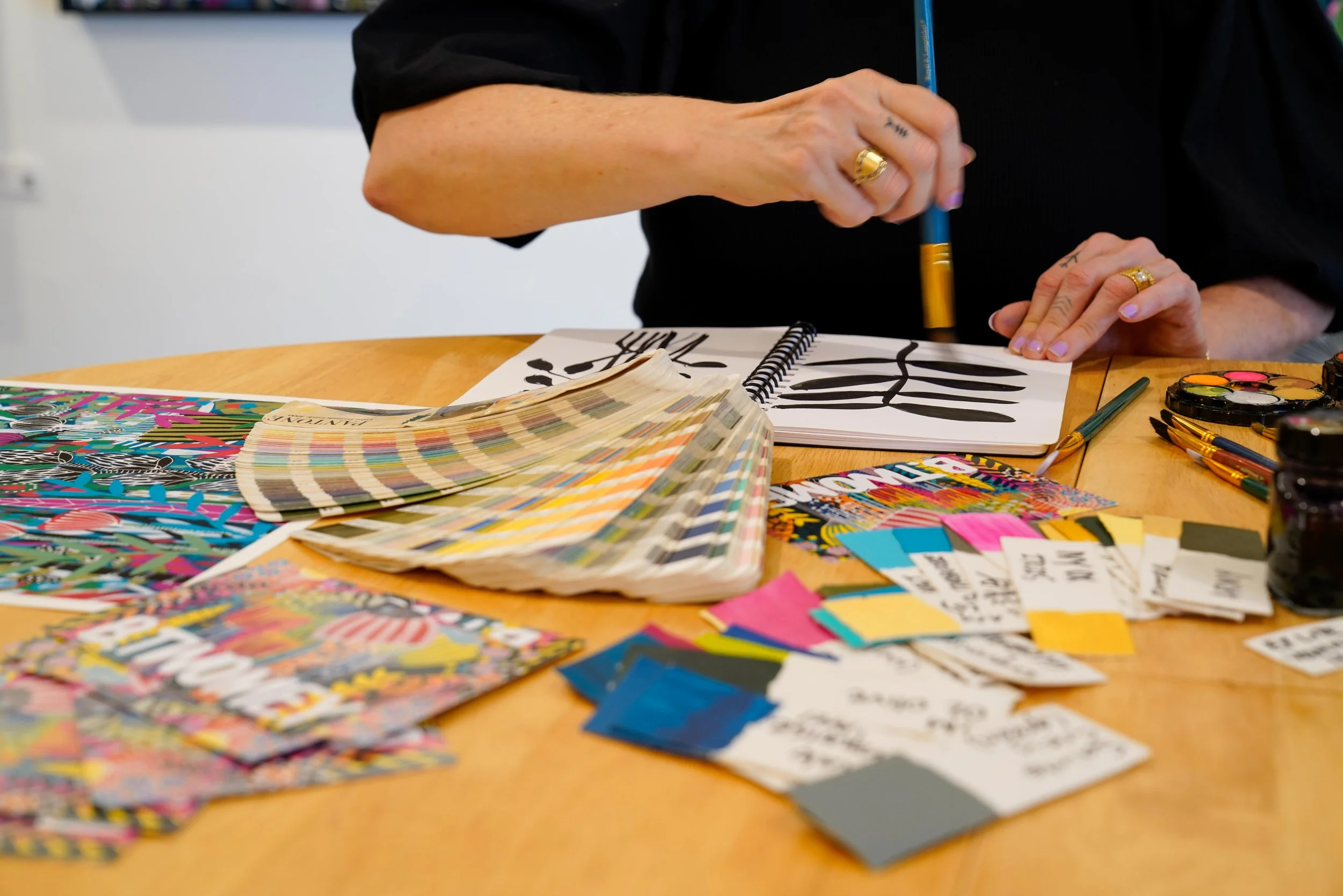 Close-up of a person's hands working on black and white abstract art, surrounded by paint swatches, color samples, and art supplies on a wooden table.
