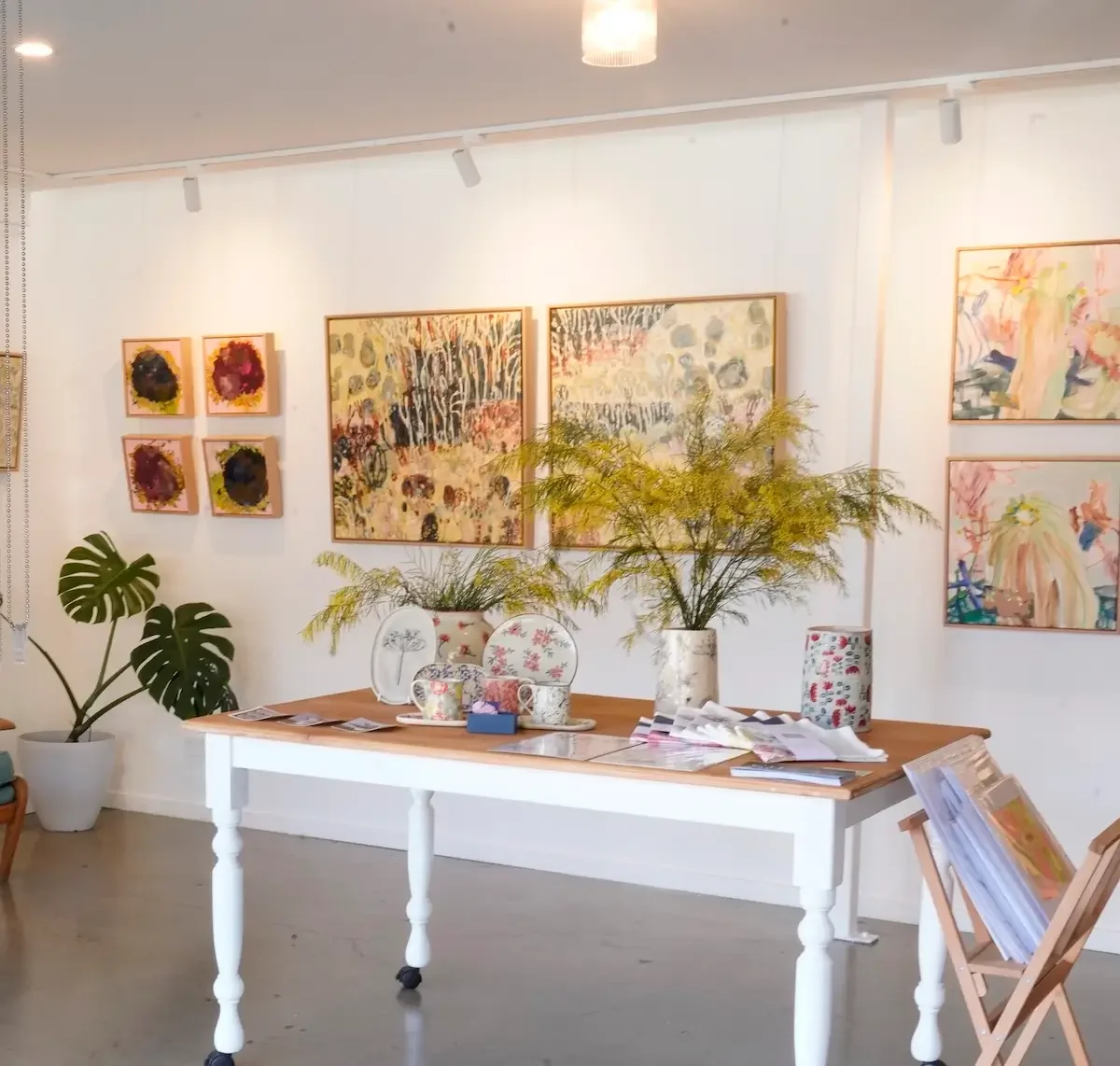 An interior scene of an art gallery or studio featuring a white table with plants, cups, and papers, surrounded by framed abstract and floral art on white walls, with a large Monstera plant in a pot on the floor to the left.