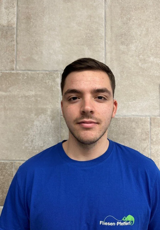 Ein junger Mann steht vor einer beige Wand, trägt ein blaues T-Shirt mit einem Logo und der Aufschrift "Fliesen-Pfeffer".