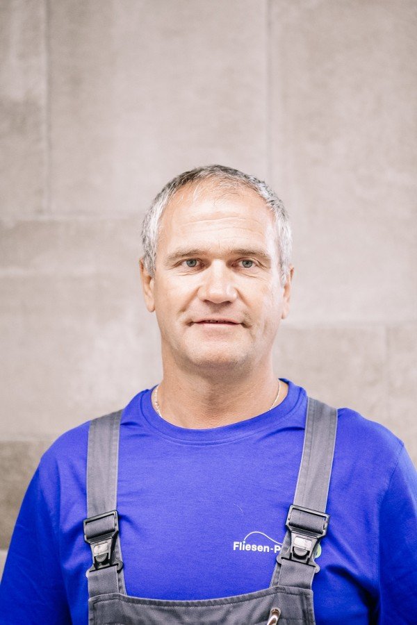 Ein Mann mit kurzen, grauen Haaren trägt eine blaue Arbeitshose und ein T-Shirt mit dem Aufdruck 'Fliesen-P' vor einer Steinwand.