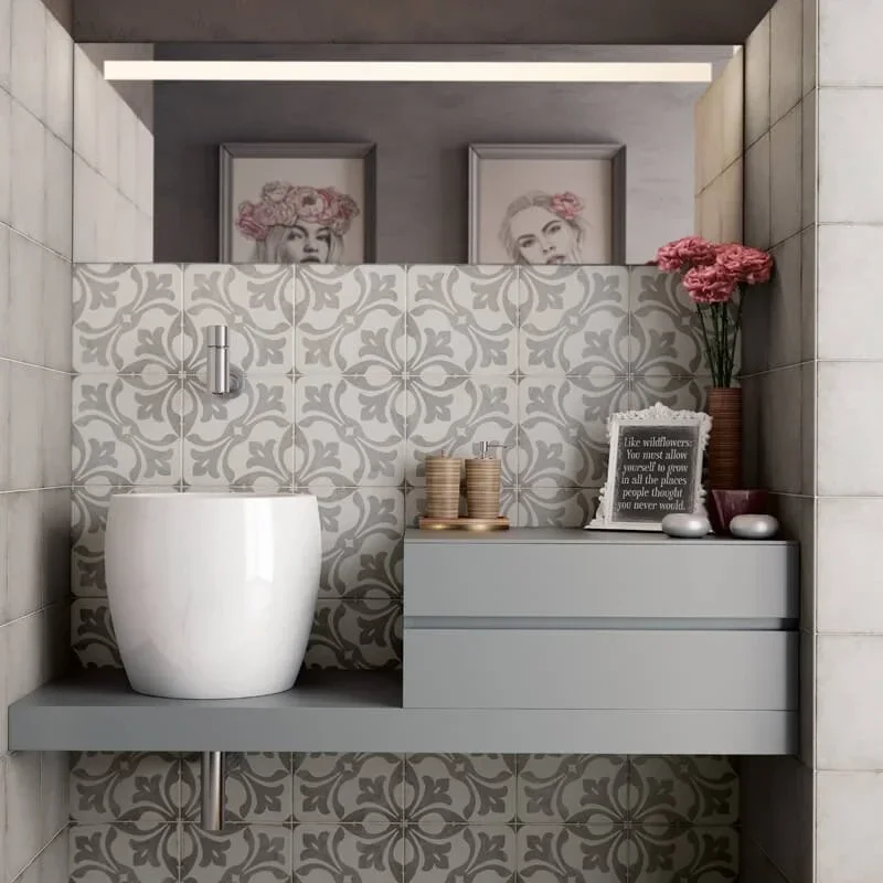Ein moderner Badezimmerbereich mit einer weißen Blumentopf-Waschschüssel, einem grauen Schrank mit Dekorationen, und gerahmten Fotos von Frauen mit Blumen im Haar im Hintergrund, dekorative Gegenstände wie Vasen, Kerzen und ein Zitat in einer kleinen Tafel.