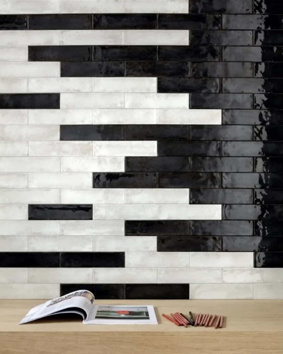 Holztisch mit einem offenen Buch, roten und schwarzen Kreiden und Bleistiften, vor einer Wand mit schwarzem und weißem Mosaikfliesen.