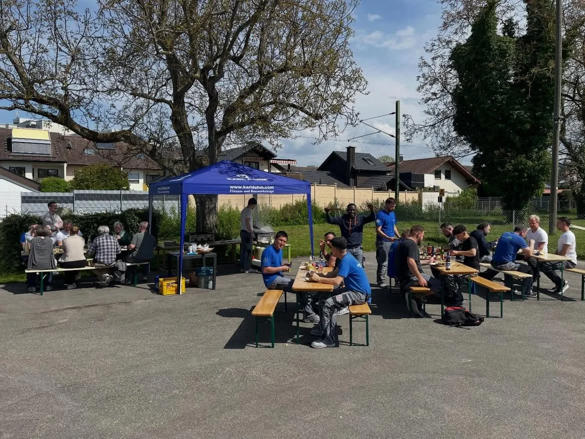 Gruppe von Menschen, die draußen an langen Tischen sitzen und essen, beim Grillen unter einem blauen Pavillon, mit Häusern im Hintergrund und einem großen Baum.